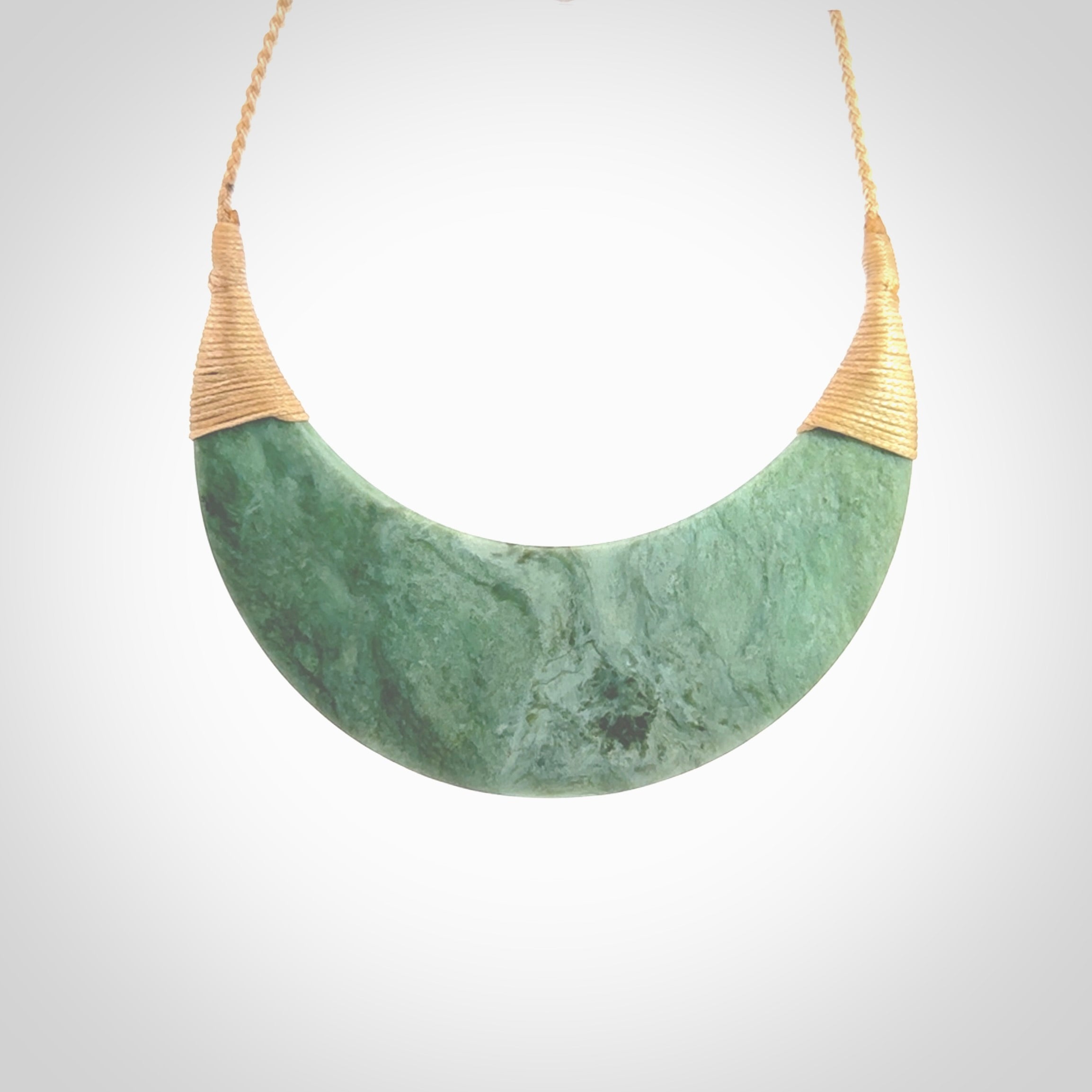 This picture shows a hand carved New Zealand Inanga jade contemporary half-shield pendant. It is a light blue and green colour and is a wonderful, large piece of jewellery. The cord is hand plaited and adjustable so that you can position the pendant where it suits you best. This piece was carved for us by Kyohei Noguchi. Delivery is free worldwide.