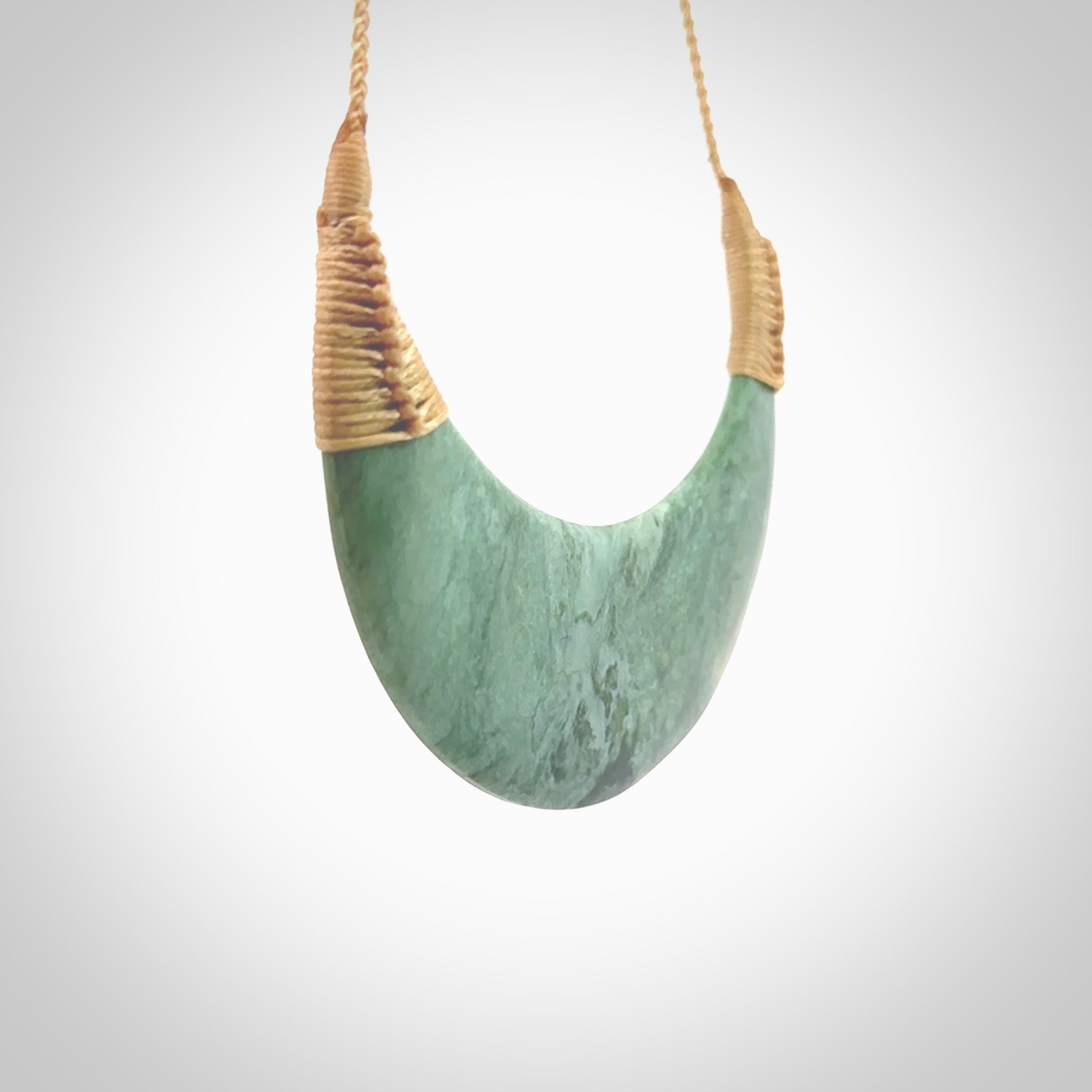 This picture shows a hand carved New Zealand Inanga jade contemporary half-shield pendant. It is a light blue and green colour and is a wonderful, large piece of jewellery. The cord is hand plaited and adjustable so that you can position the pendant where it suits you best. This piece was carved for us by Kyohei Noguchi. Delivery is free worldwide.