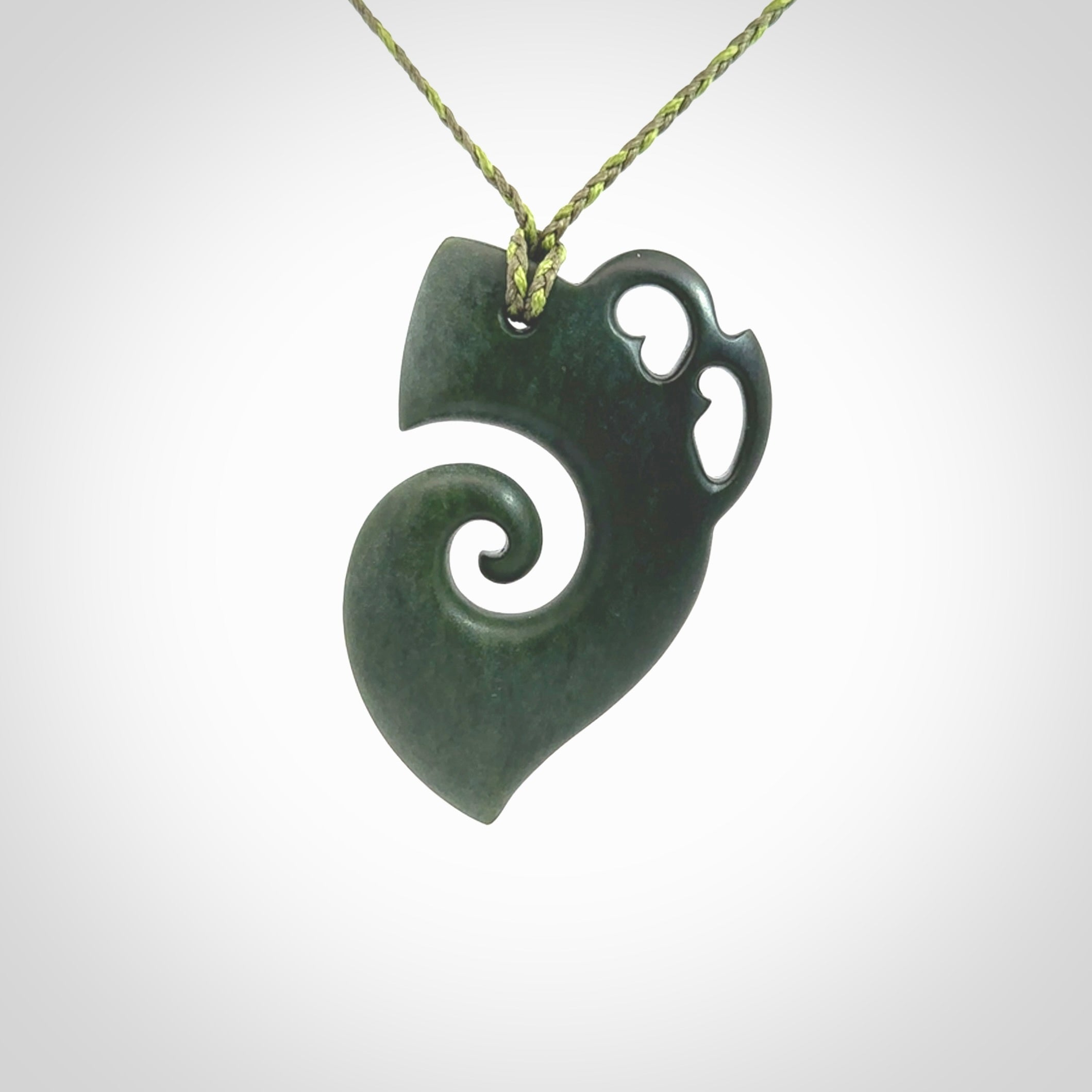 This photo shows a koru with a Kea head carved into the leading edge. The artist, Kerry Thompson, has carved this beautifully and he has polished parts of the carving to a high shine, and other parts he has left in a matte finish. The contrast is beautiful. The pendant is suspended from a plaited grass green/sage green cord which is adjustable.