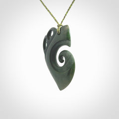 This photo shows a koru with a Kea head carved into the leading edge. The artist, Kerry Thompson, has carved this beautifully and he has polished parts of the carving to a high shine, and other parts he has left in a matte finish. The contrast is beautiful. The pendant is suspended from a plaited grass green/sage green cord which is adjustable.