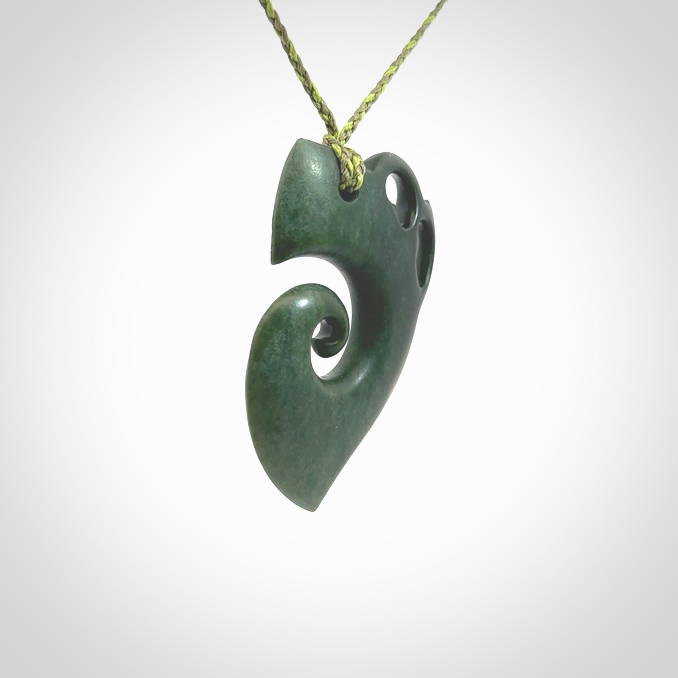 This photo shows a koru with a Kea head carved into the leading edge. The artist, Kerry Thompson, has carved this beautifully and he has polished parts of the carving to a high shine, and other parts he has left in a matte finish. The contrast is beautiful. The pendant is suspended from a plaited grass green/sage green cord which is adjustable.