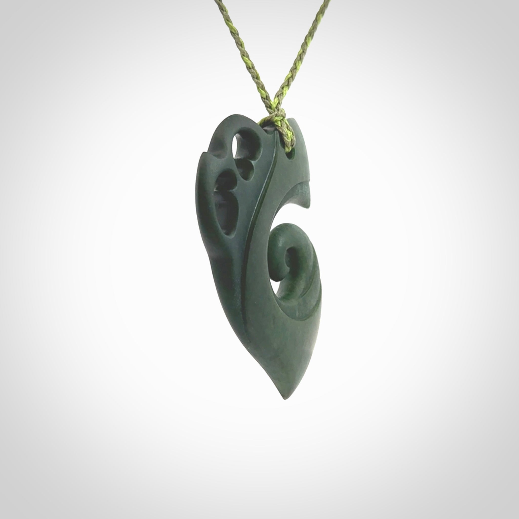 This photo shows a koru with a Kea head carved into the leading edge. The artist, Kerry Thompson, has carved this beautifully and he has polished parts of the carving to a high shine, and other parts he has left in a matte finish. The contrast is beautiful. The pendant is suspended from a plaited grass green/sage green cord which is adjustable.