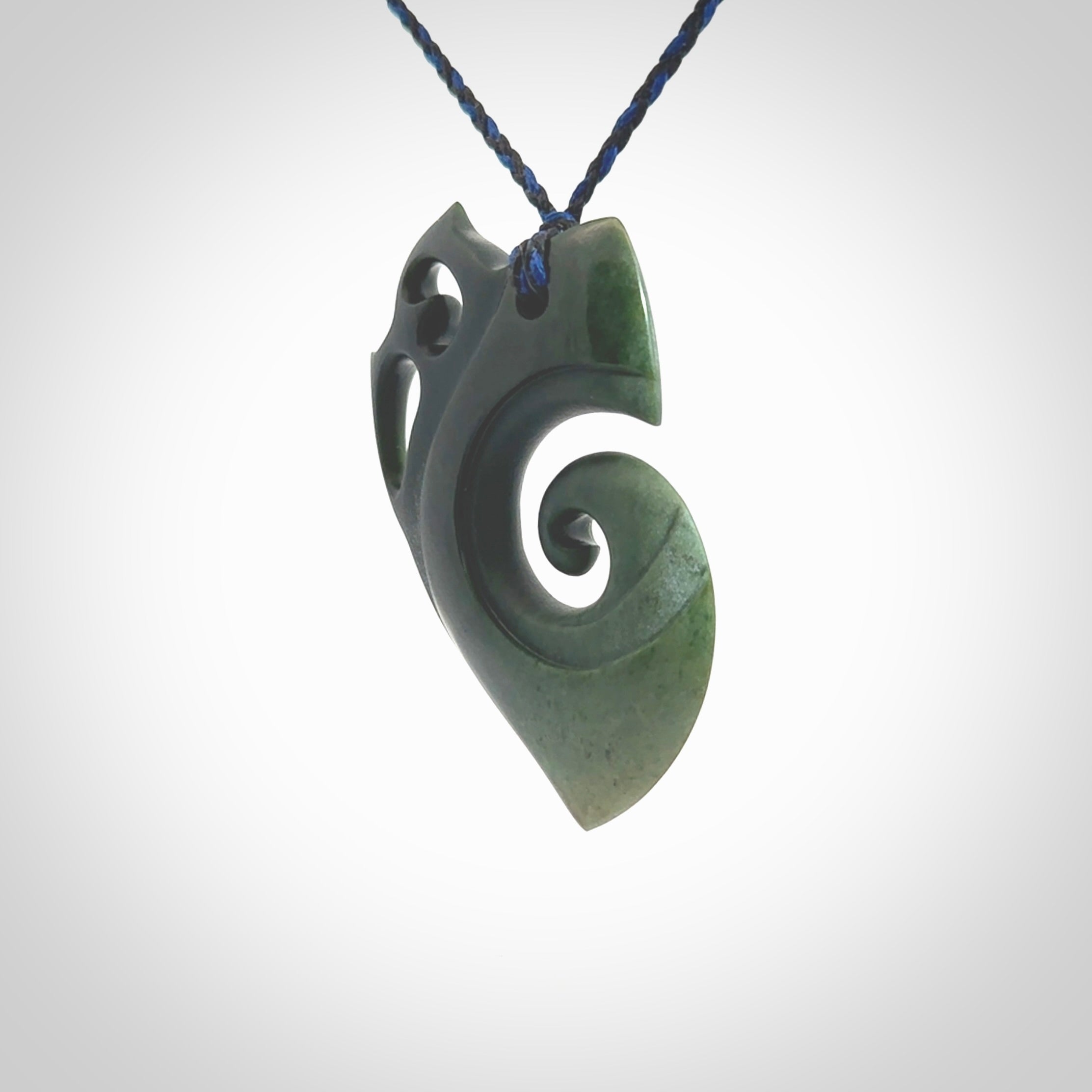 This photo shows a koru with a Owl head carved into the leading edge. The artist, Kerry Thompson, has carved this beautifully and he has polished parts of the carving to a high shine, and other parts he has left in a matte finish. The contrast is beautiful. The pendant is suspended from a plaited pacific blue/black cord which is adjustable.