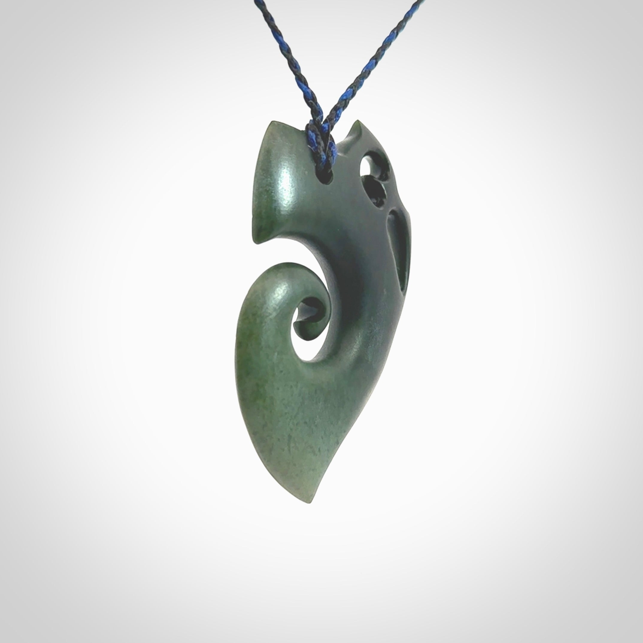 This photo shows a koru with a Owl head carved into the leading edge. The artist, Kerry Thompson, has carved this beautifully and he has polished parts of the carving to a high shine, and other parts he has left in a matte finish. The contrast is beautiful. The pendant is suspended from a plaited pacific blue/black cord which is adjustable.