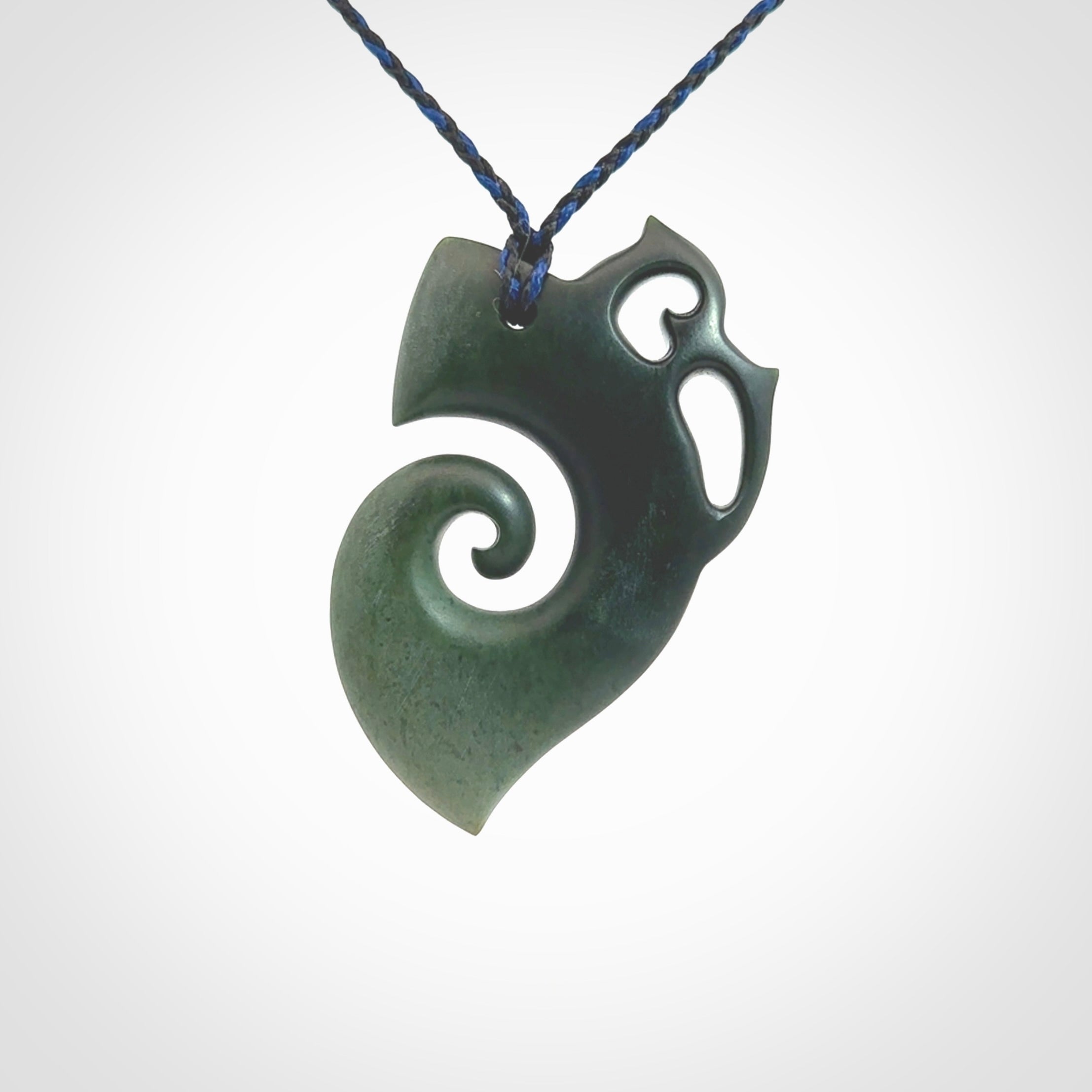 This photo shows a koru with a Owl head carved into the leading edge. The artist, Kerry Thompson, has carved this beautifully and he has polished parts of the carving to a high shine, and other parts he has left in a matte finish. The contrast is beautiful. The pendant is suspended from a plaited pacific blue/black cord which is adjustable.