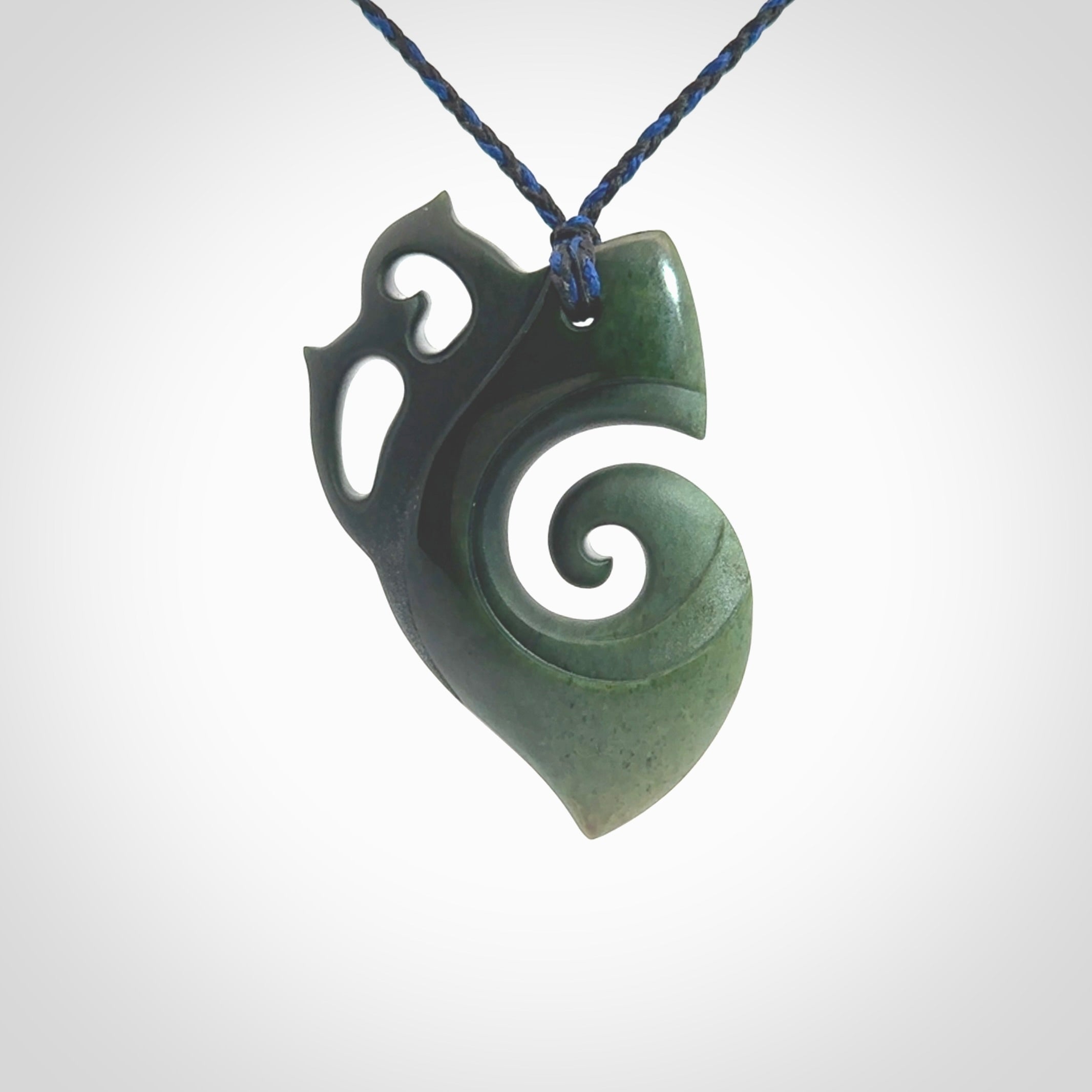 This photo shows a koru with a Owl head carved into the leading edge. The artist, Kerry Thompson, has carved this beautifully and he has polished parts of the carving to a high shine, and other parts he has left in a matte finish. The contrast is beautiful. The pendant is suspended from a plaited pacific blue/black cord which is adjustable.