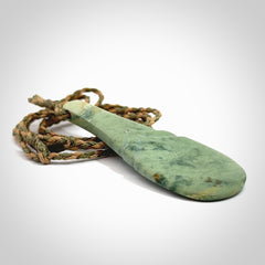 This is a sleek and slender hand carved New Zealand Jade mere pendant. The stone is a very pale green with mottled pale inclusions. The cord is tan and khaki and adjustable. The pendant is delivered in a woven kete pouch.