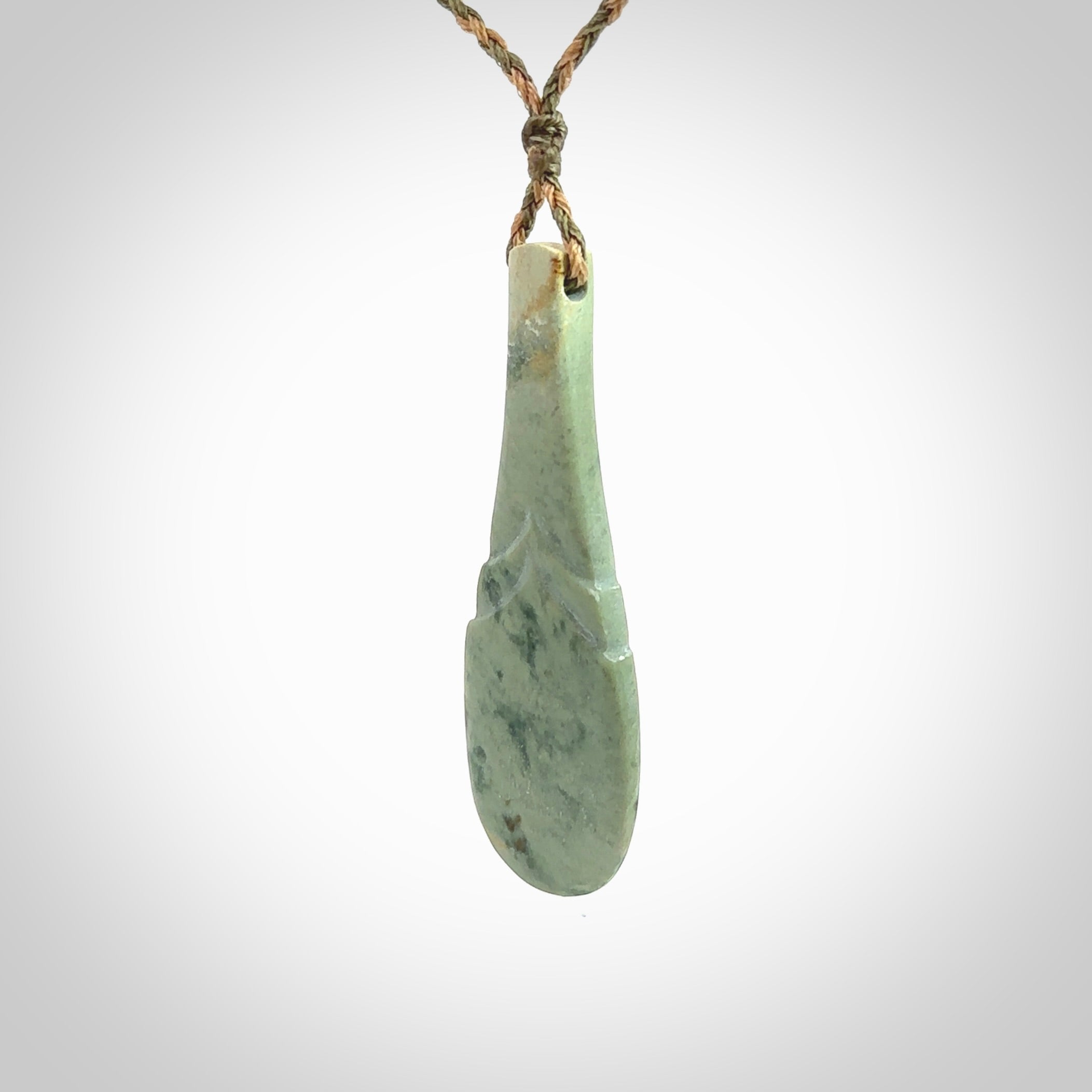 This is a sleek and slender hand carved New Zealand Jade mere pendant. The stone is a very pale green with mottled pale inclusions. The cord is tan and khaki and adjustable. The pendant is delivered in a woven kete pouch.