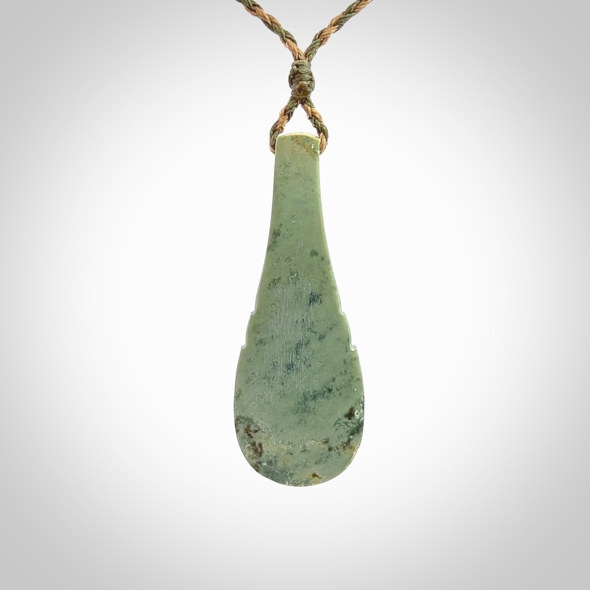 This is a sleek and slender hand carved New Zealand Jade mere pendant. The stone is a very pale green with mottled pale inclusions. The cord is tan and khaki and adjustable. The pendant is delivered in a woven kete pouch.