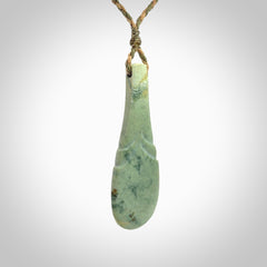 This is a sleek and slender hand carved New Zealand Jade mere pendant. The stone is a very pale green with mottled pale inclusions. The cord is tan and khaki and adjustable. The pendant is delivered in a woven kete pouch.