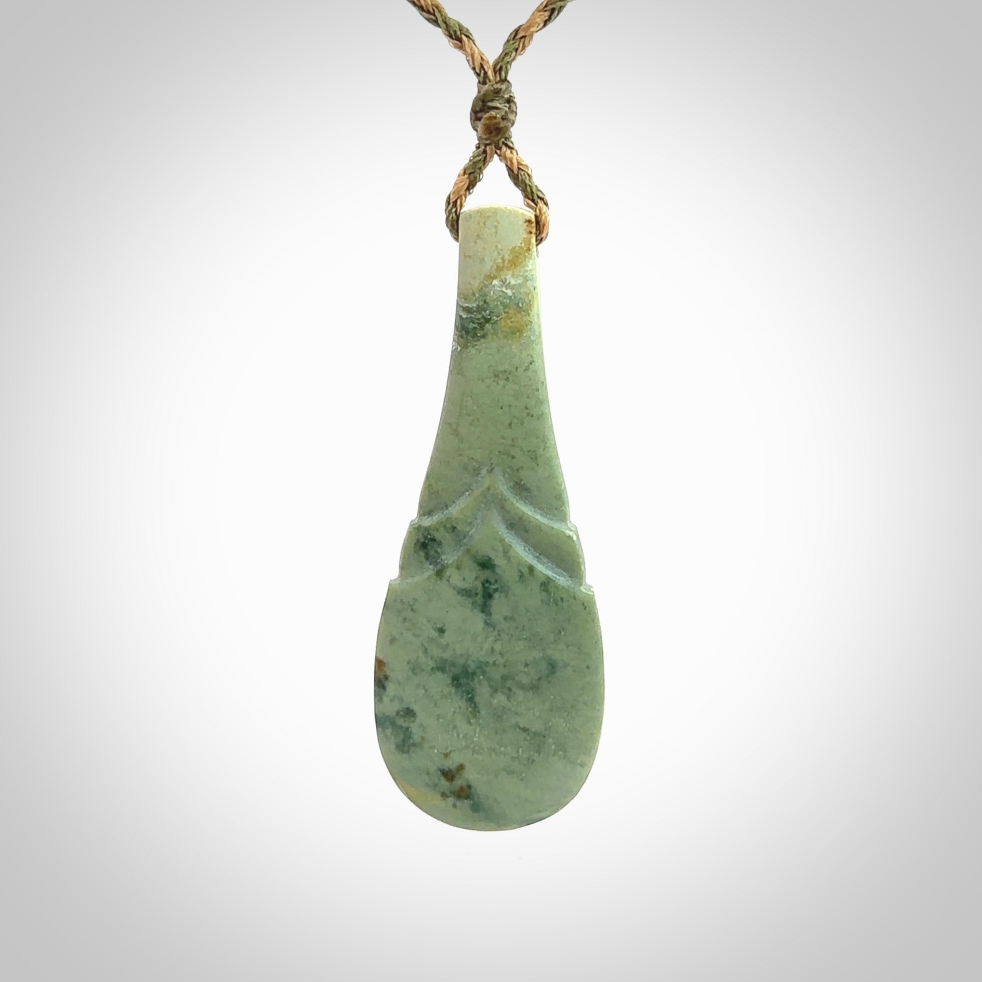 This is a sleek and slender hand carved New Zealand Jade mere pendant. The stone is a very pale green with mottled pale inclusions. The cord is tan and khaki and adjustable. The pendant is delivered in a woven kete pouch.