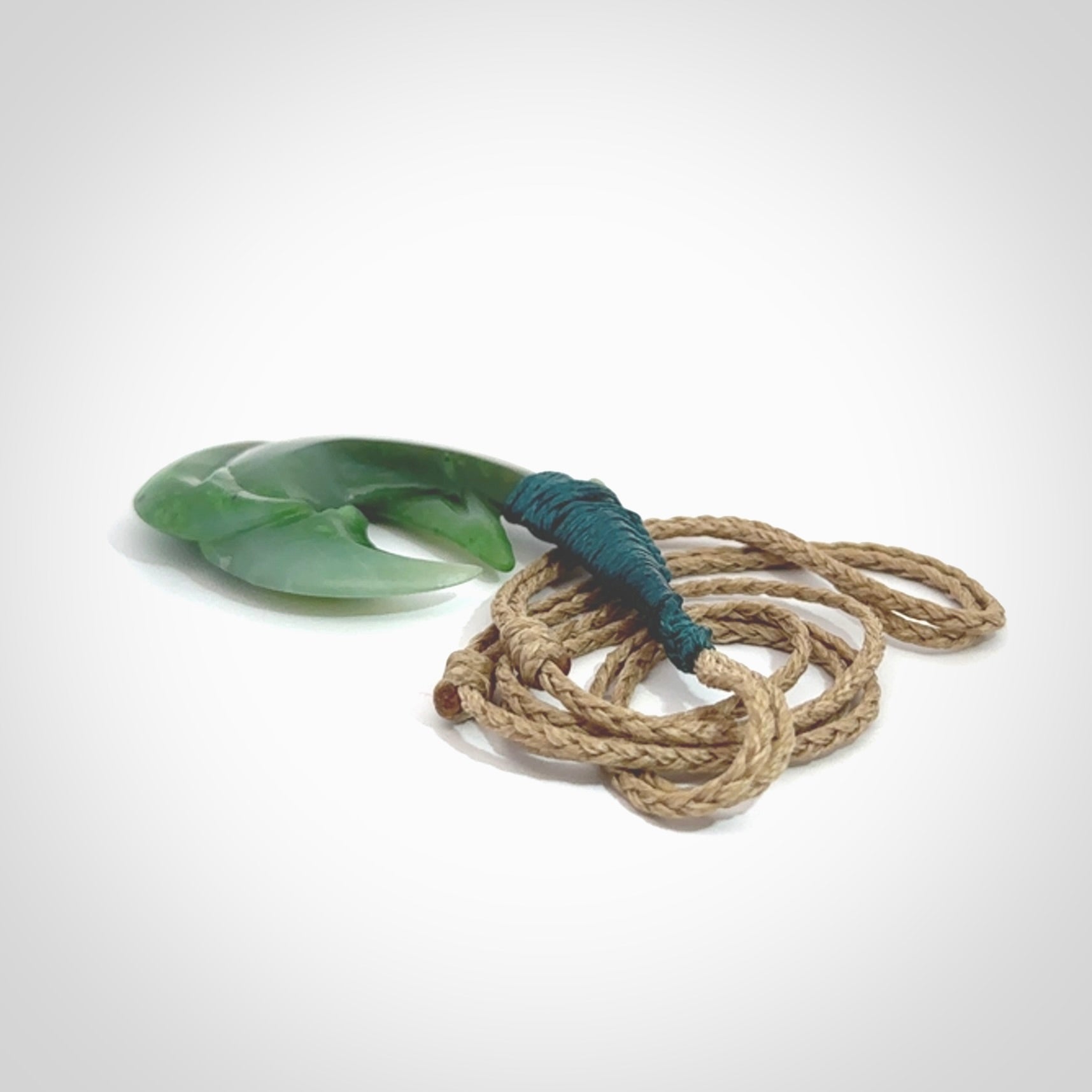 Hand carved Jade matau hook pendant. This picture shows the pendant with a Kalahari Tan cord and a Sea Moss Blue binding. The cord is length adjustable. It is a double hook with a larger hook and a smaller one wrapped around the bottom of the larger piece. This pendant has been designed for both men and women to wear. NZ Pacific art to wear.