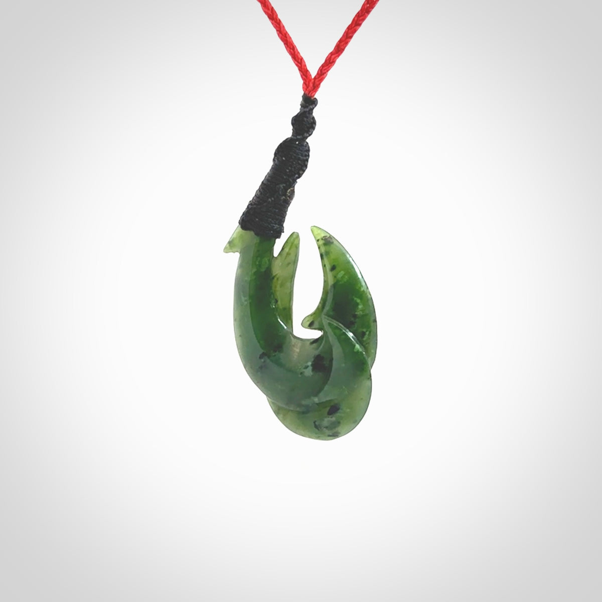 Hand carved Jade matau hook pendant. This picture shows the pendant with a red cord and a black binding. The cord is length adjustable. It is a double hook with a larger hook and a smaller one wrapped around the bottom of the larger piece. This pendant has been designed for both men and women to wear. NZ Pacific art to wear.