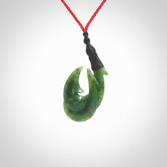 Hand carved Jade matau hook pendant. This picture shows the pendant with a red cord and a black binding. The cord is length adjustable. It is a double hook with a larger hook and a smaller one wrapped around the bottom of the larger piece. This pendant has been designed for both men and women to wear. NZ Pacific art to wear.