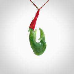 Hand carved Jade matau hook pendant. This picture shows the pendant with a red/khaki cord and a red binding. The cord is length adjustable. It is a double hook with a larger hook and a smaller one wrapped around the bottom of the larger piece. This pendant has been designed for both men and women to wear. NZ Pacific art to wear.