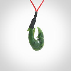 Hand carved Jade matau hook pendant. This picture shows the pendant with a red cord and a black binding. The cord is length adjustable. It is a double hook with a larger hook and a smaller one wrapped around the bottom of the larger piece. This pendant has been designed for both men and women to wear. NZ Pacific art to wear.