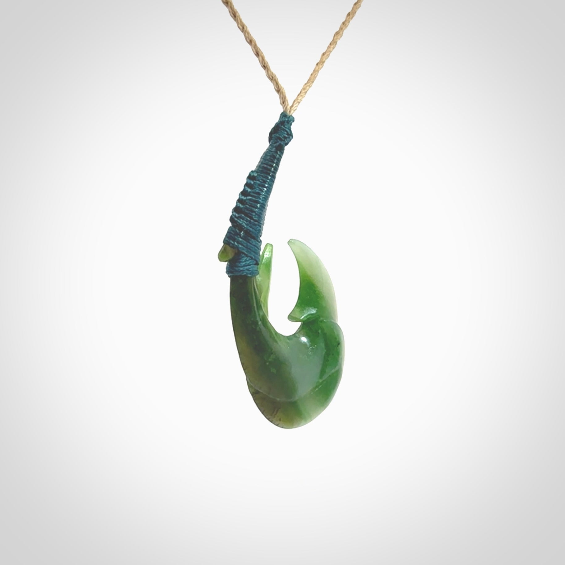 Hand carved Jade matau hook pendant. This picture shows the pendant with a Kalahari Tan cord and a Sea Moss Blue binding. The cord is length adjustable. It is a double hook with a larger hook and a smaller one wrapped around the bottom of the larger piece. This pendant has been designed for both men and women to wear. NZ Pacific art to wear.