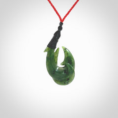 Hand carved Jade matau hook pendant. This picture shows the pendant with a red cord and a black binding. The cord is length adjustable. It is a double hook with a larger hook and a smaller one wrapped around the bottom of the larger piece. This pendant has been designed for both men and women to wear. NZ Pacific art to wear.