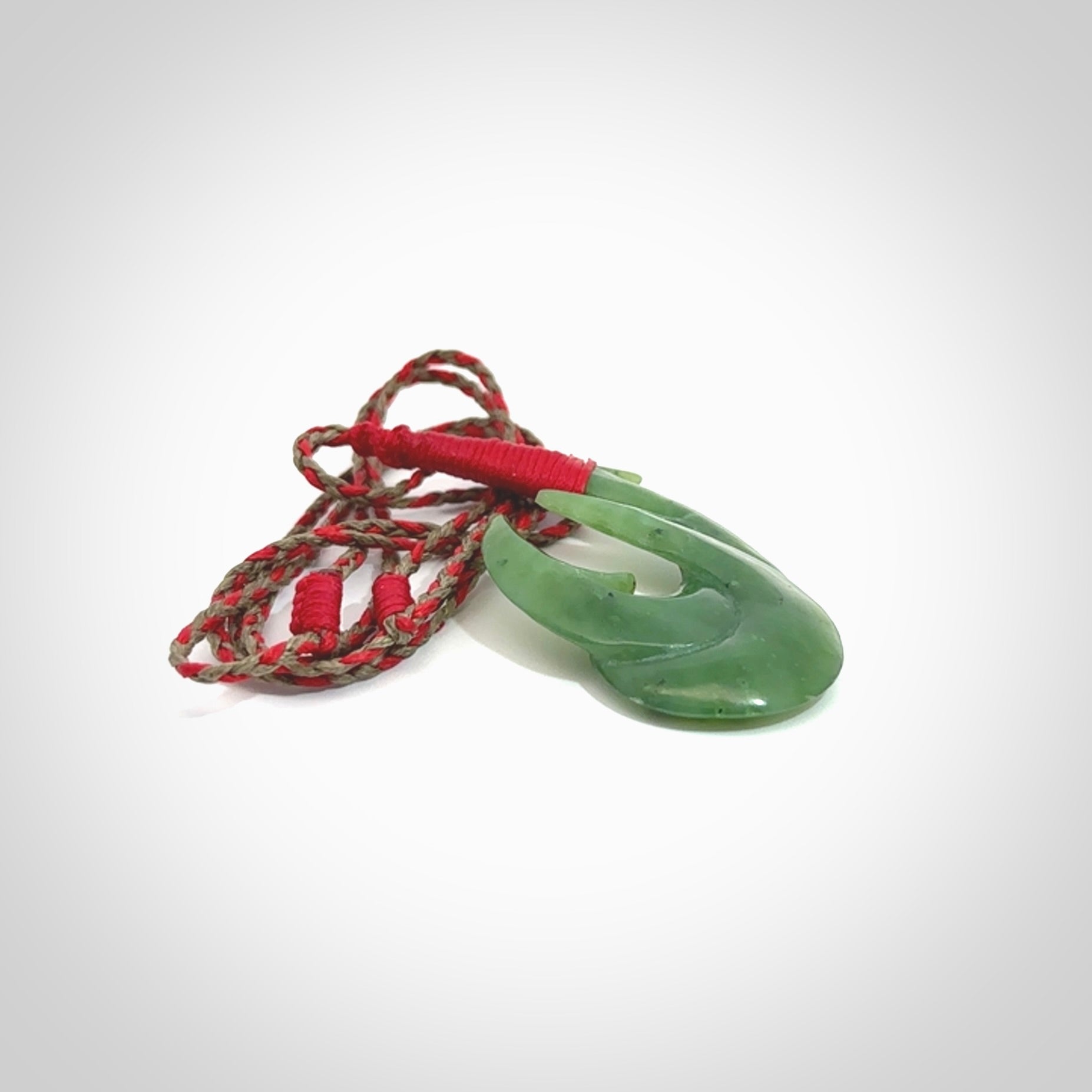 Hand carved Jade matau hook pendant. This picture shows the pendant with a red/khaki cord and a red binding. The cord is length adjustable. It is a double hook with a larger hook and a smaller one wrapped around the bottom of the larger piece. This pendant has been designed for both men and women to wear. NZ Pacific art to wear.