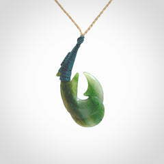 Hand carved Jade matau hook pendant. This picture shows the pendant with a Kalahari Tan cord and a Sea Moss Blue binding. The cord is length adjustable. It is a double hook with a larger hook and a smaller one wrapped around the bottom of the larger piece. This pendant has been designed for both men and women to wear. NZ Pacific art to wear.