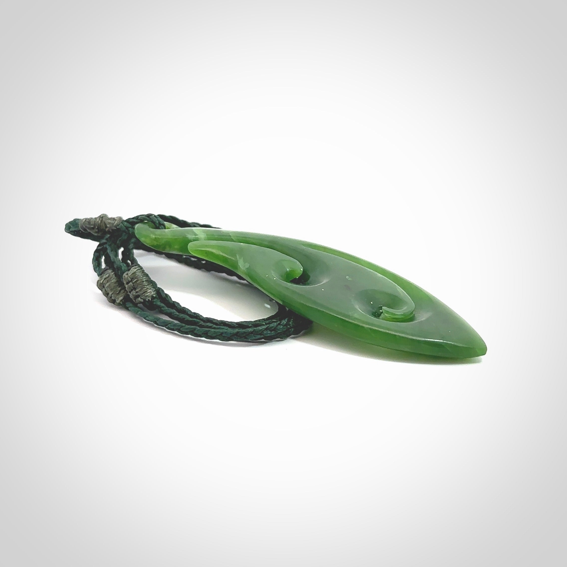 This photo shows a large matau, or hook, pendant carved in deep green New Zealand Jade. It is a very interesting piece which has a koru carved into the centre of the hook. It is strung on a manuka green cord which is adjustable. The artist who carved this pendant is Ross Crump, a master carver of jade from New Zealand.