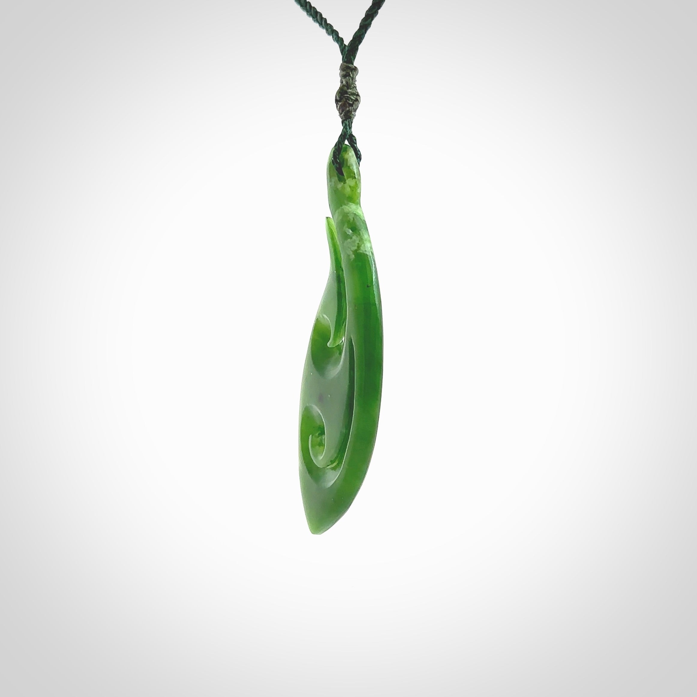 This photo shows a large matau, or hook, pendant carved in deep green New Zealand Jade. It is a very interesting piece which has a koru carved into the centre of the hook. It is strung on a manuka green cord which is adjustable. The artist who carved this pendant is Ross Crump, a master carver of jade from New Zealand.