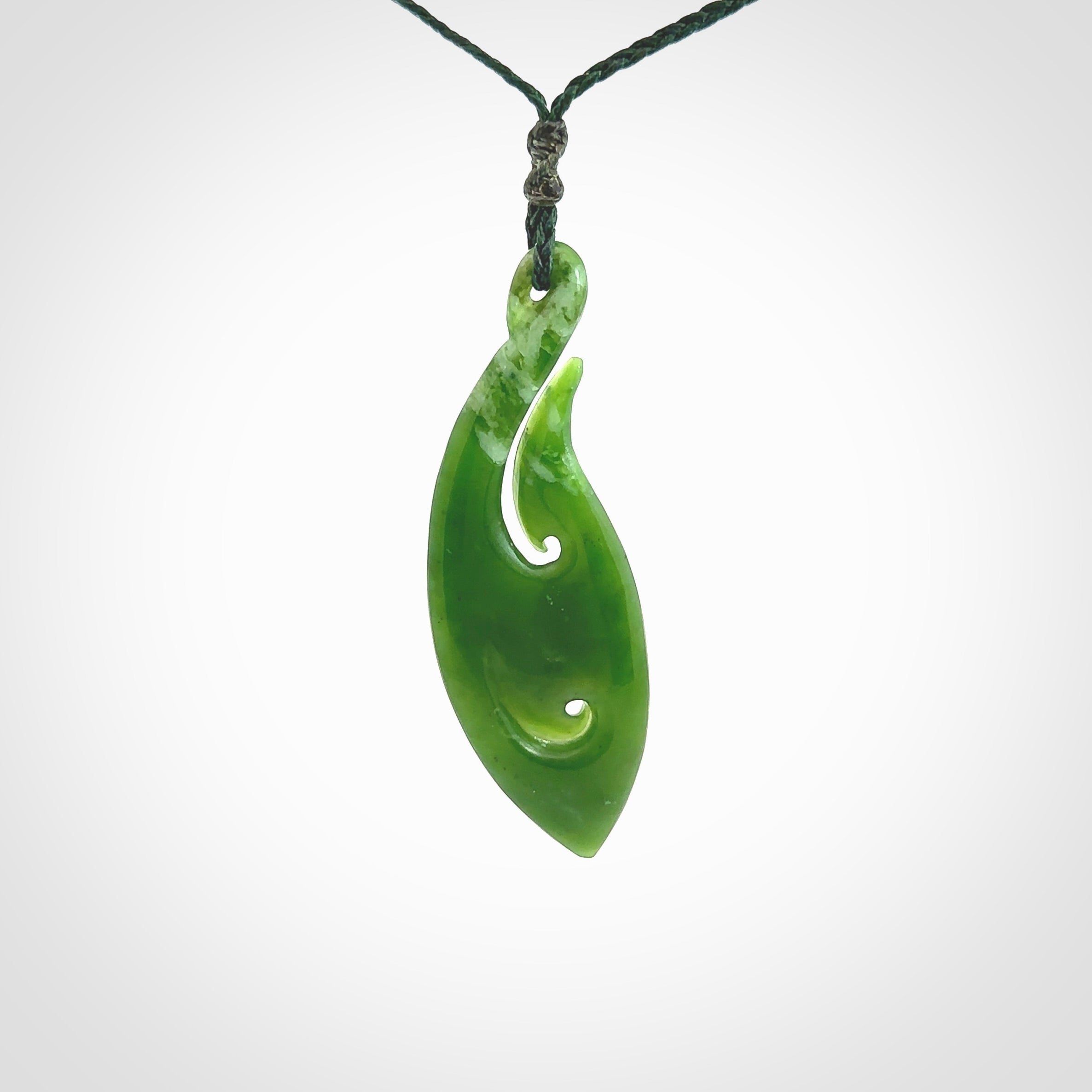 This photo shows a large matau, or hook, pendant carved in deep green New Zealand Jade. It is a very interesting piece which has a koru carved into the centre of the hook. It is strung on a manuka green cord which is adjustable. The artist who carved this pendant is Ross Crump, a master carver of jade from New Zealand.