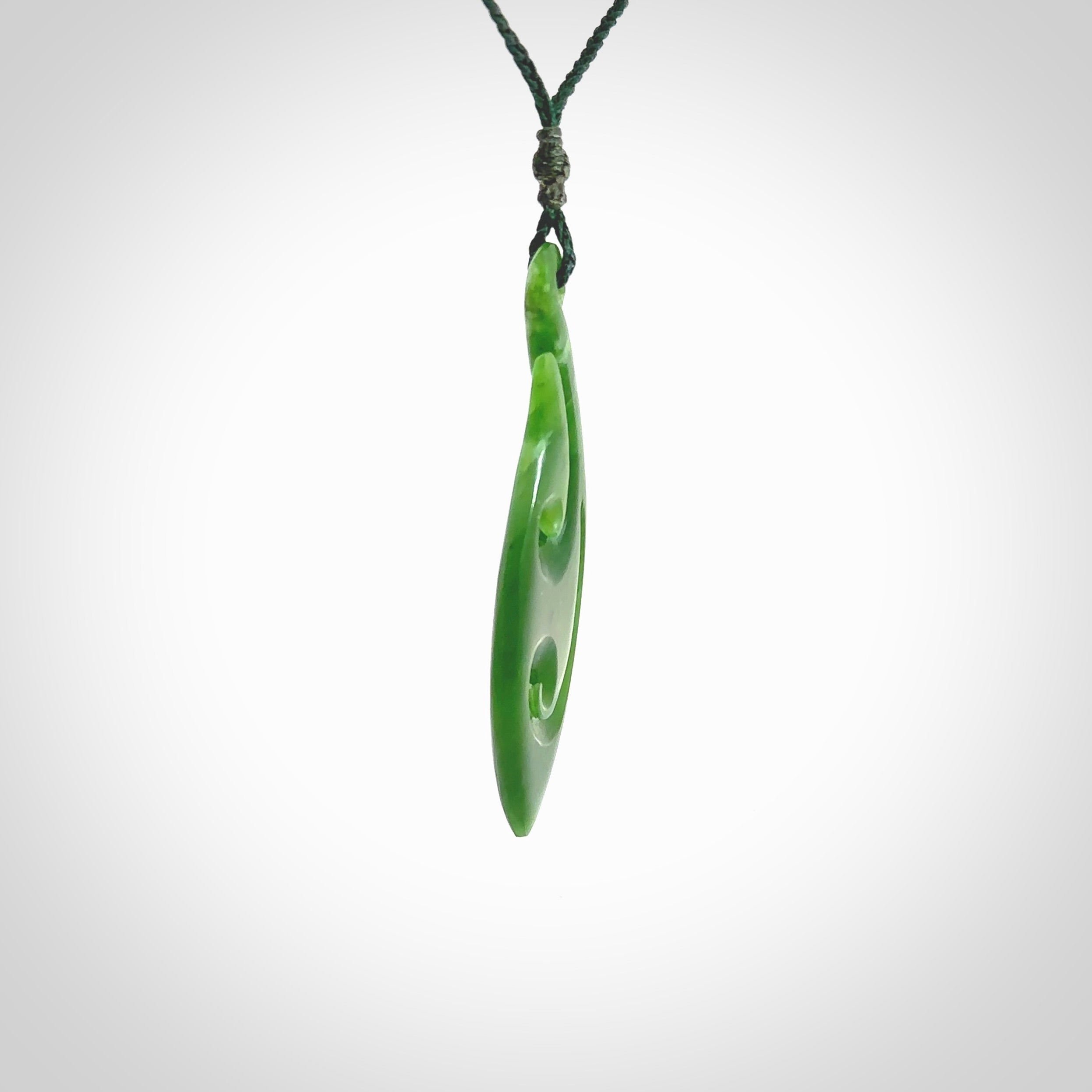 This photo shows a large matau, or hook, pendant carved in deep green New Zealand Jade. It is a very interesting piece which has a koru carved into the centre of the hook. It is strung on a manuka green cord which is adjustable. The artist who carved this pendant is Ross Crump, a master carver of jade from New Zealand.