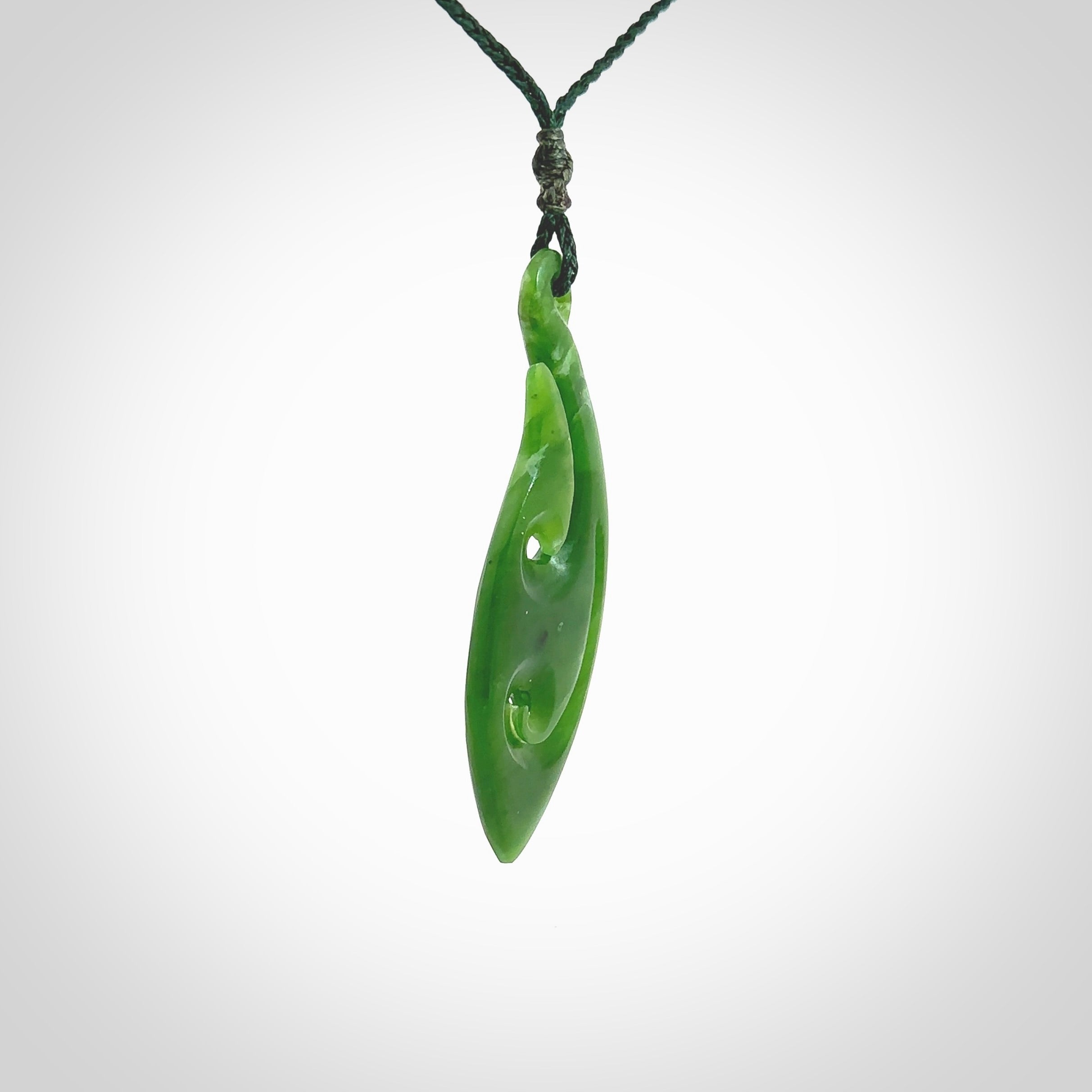 This photo shows a large matau, or hook, pendant carved in deep green New Zealand Jade. It is a very interesting piece which has a koru carved into the centre of the hook. It is strung on a manuka green cord which is adjustable. The artist who carved this pendant is Ross Crump, a master carver of jade from New Zealand.