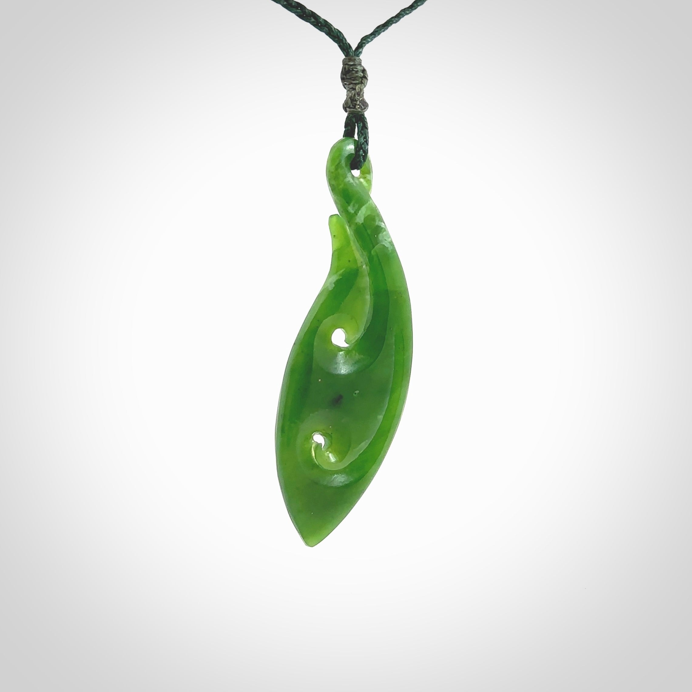 This photo shows a large matau, or hook, pendant carved in deep green New Zealand Jade. It is a very interesting piece which has a koru carved into the centre of the hook. It is strung on a manuka green cord which is adjustable. The artist who carved this pendant is Ross Crump, a master carver of jade from New Zealand.