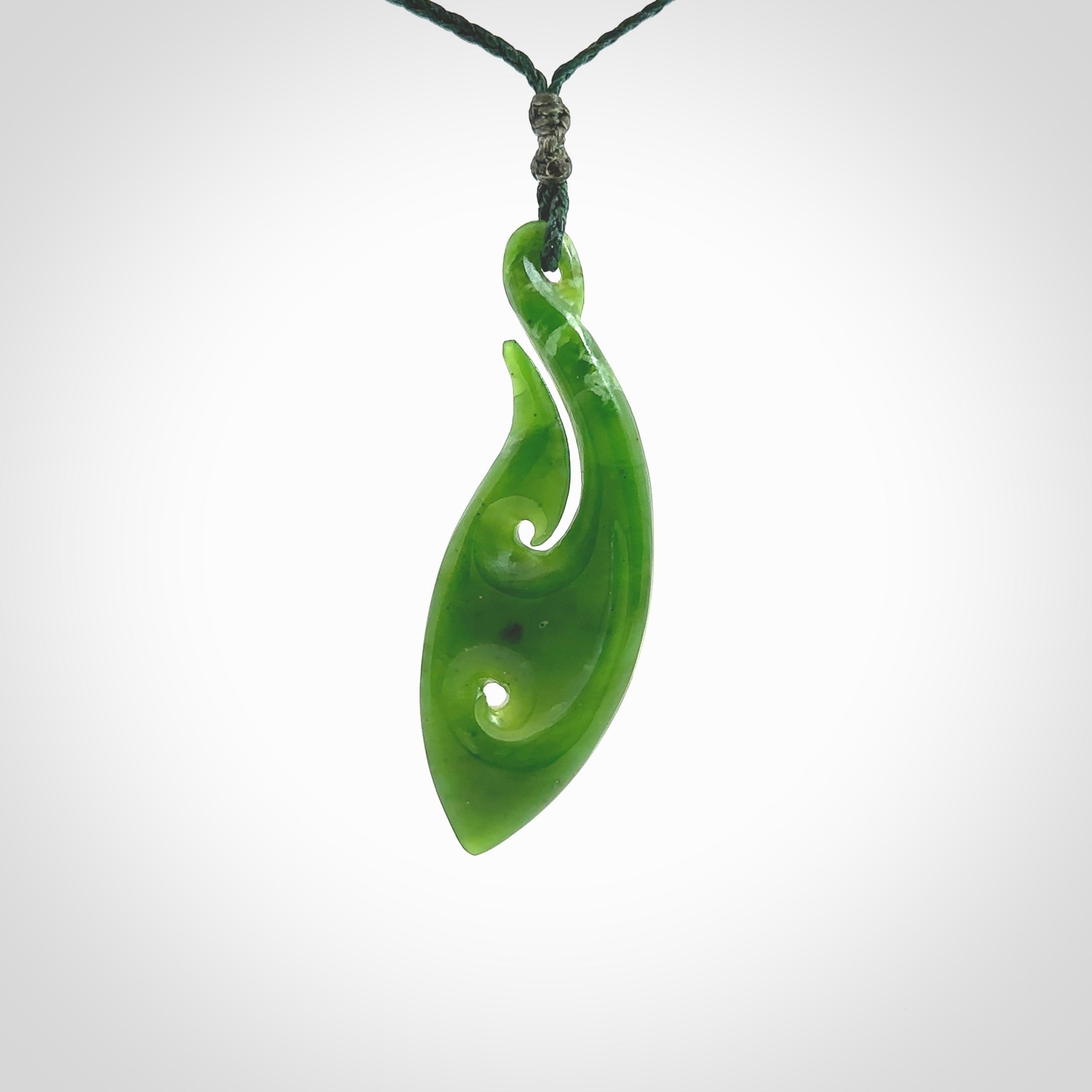 This photo shows a large matau, or hook, pendant carved in deep green New Zealand Jade. It is a very interesting piece which has a koru carved into the centre of the hook. It is strung on a manuka green cord which is adjustable. The artist who carved this pendant is Ross Crump, a master carver of jade from New Zealand.