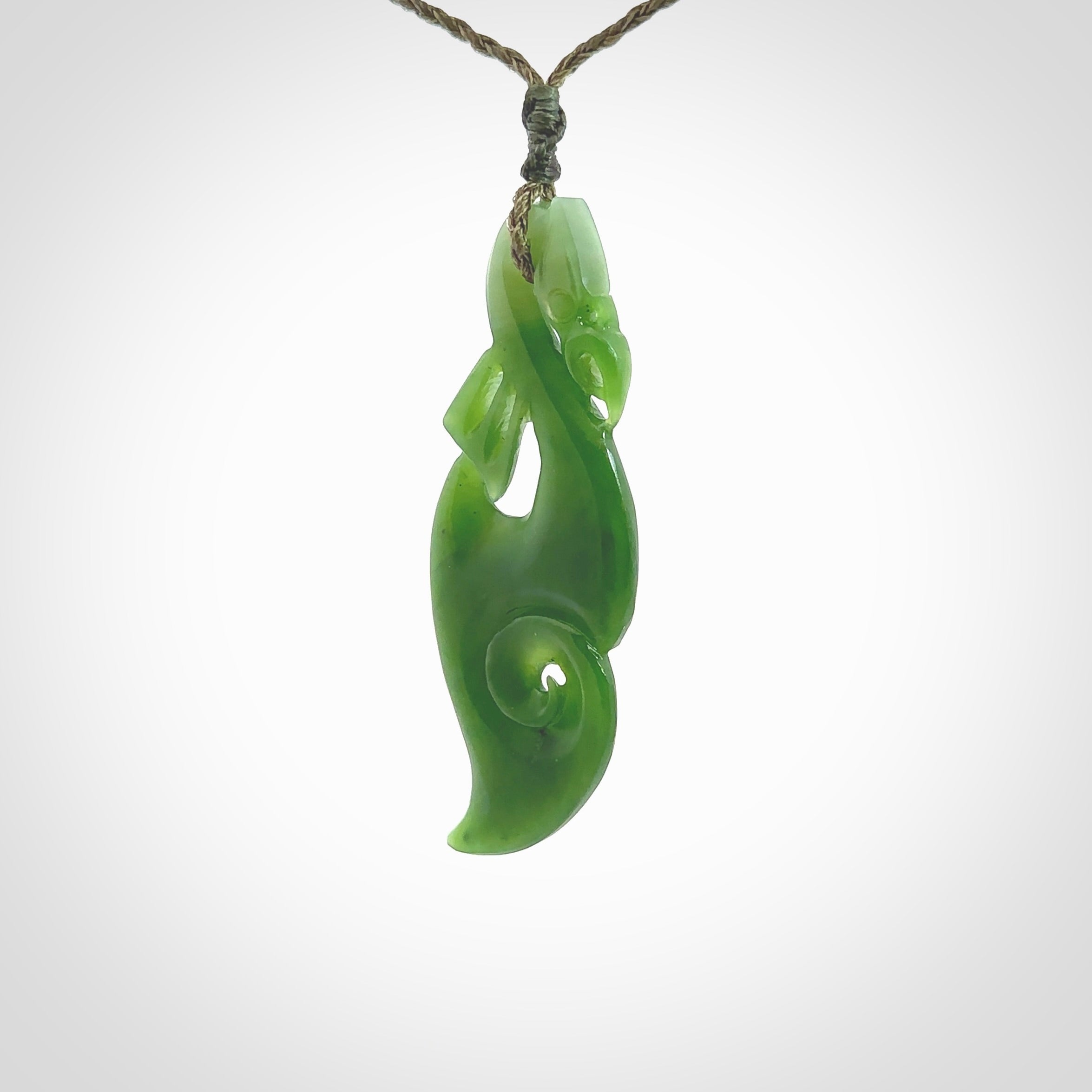 This picture shows a polished manaia pendant carved for us by Ross Crump. It is a dark matte green colour in traditional manaia design. Ross has added a koru on the bottom which elongates the carving and gives it some decorative flair. We have added a 4-Plait cord in olive green and made it length adjustable. This is a very beautiful piece of art to wear.