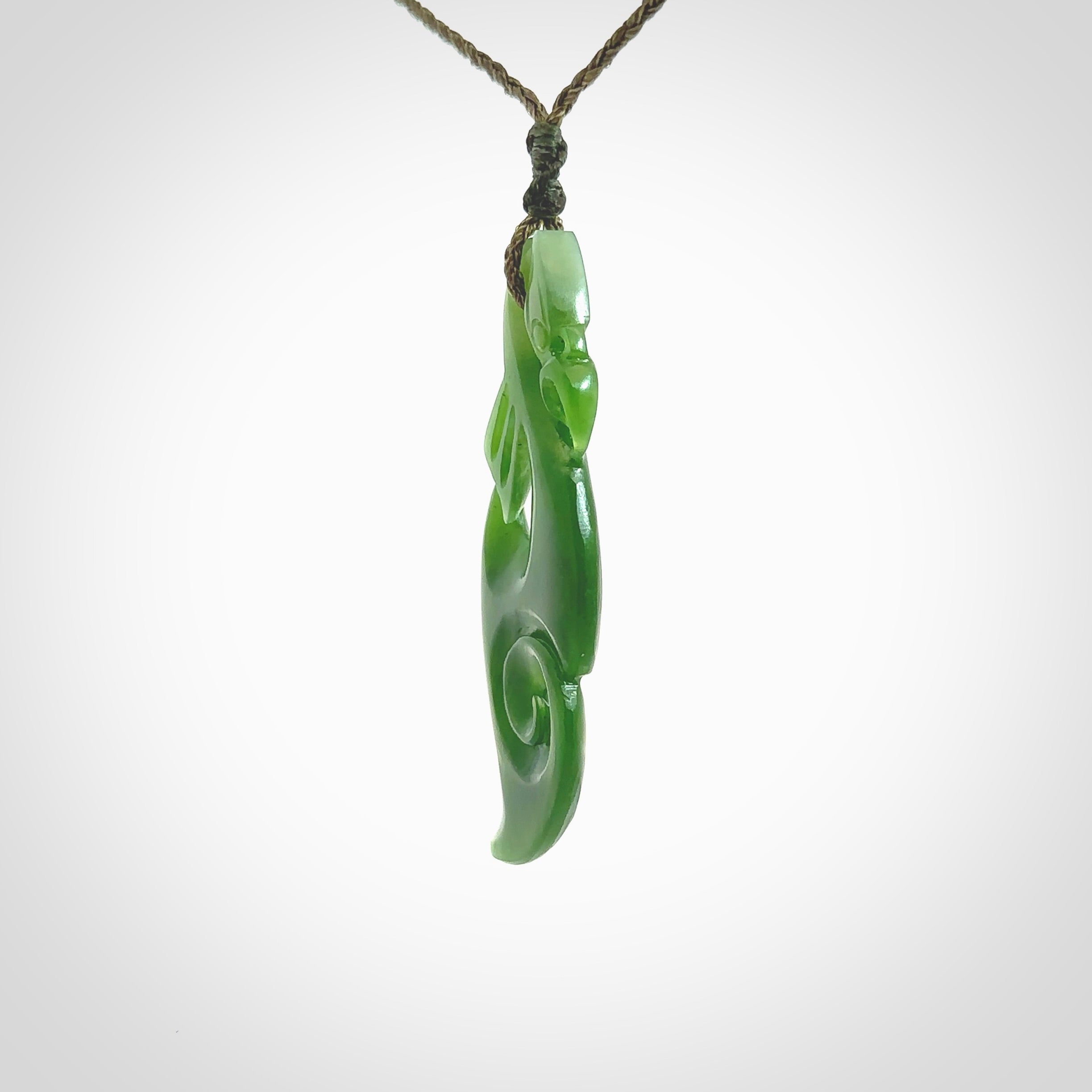 This picture shows a polished manaia pendant carved for us by Ross Crump. It is a dark matte green colour in traditional manaia design. Ross has added a koru on the bottom which elongates the carving and gives it some decorative flair. We have added a 4-Plait cord in olive green and made it length adjustable. This is a very beautiful piece of art to wear.