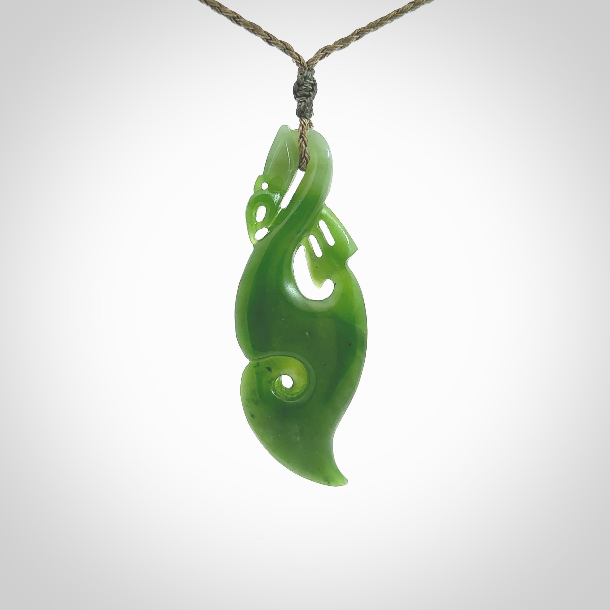 This picture shows a polished manaia pendant carved for us by Ross Crump. It is a dark matte green colour in traditional manaia design. Ross has added a koru on the bottom which elongates the carving and gives it some decorative flair. We have added a 4-Plait cord in olive green and made it length adjustable. This is a very beautiful piece of art to wear.