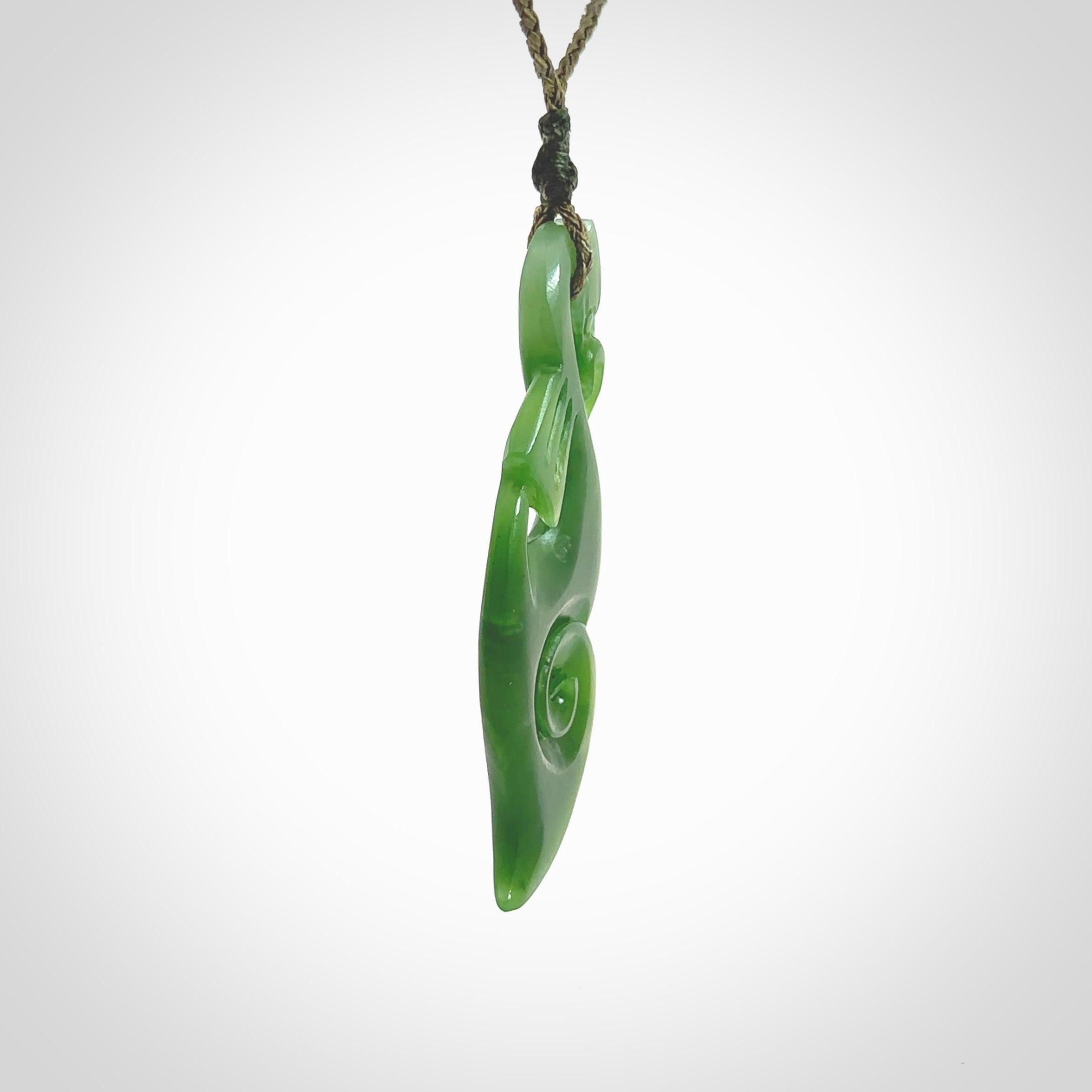 This picture shows a polished manaia pendant carved for us by Ross Crump. It is a dark matte green colour in traditional manaia design. Ross has added a koru on the bottom which elongates the carving and gives it some decorative flair. We have added a 4-Plait cord in olive green and made it length adjustable. This is a very beautiful piece of art to wear.