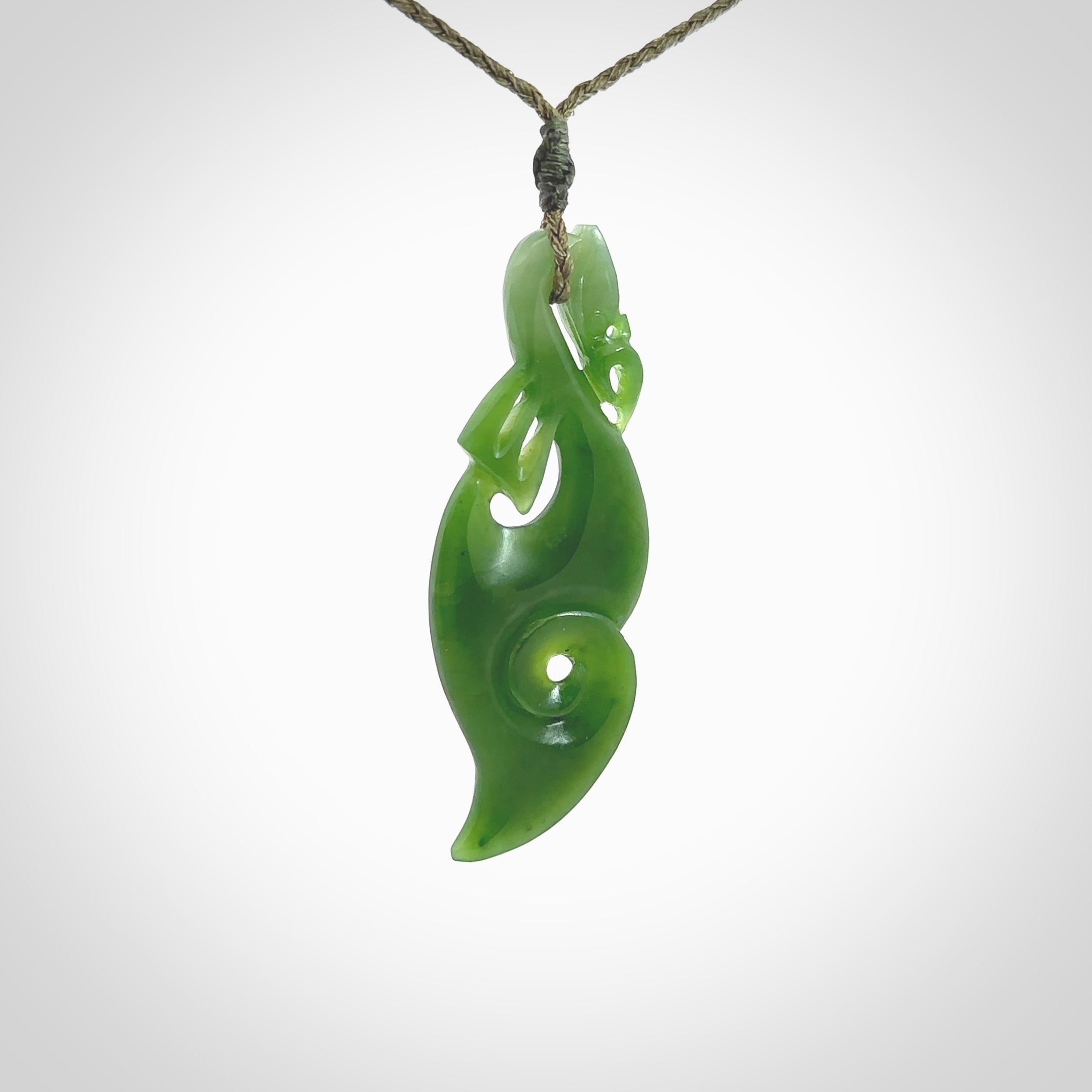 This picture shows a polished manaia pendant carved for us by Ross Crump. It is a dark matte green colour in traditional manaia design. Ross has added a koru on the bottom which elongates the carving and gives it some decorative flair. We have added a 4-Plait cord in olive green and made it length adjustable. This is a very beautiful piece of art to wear.