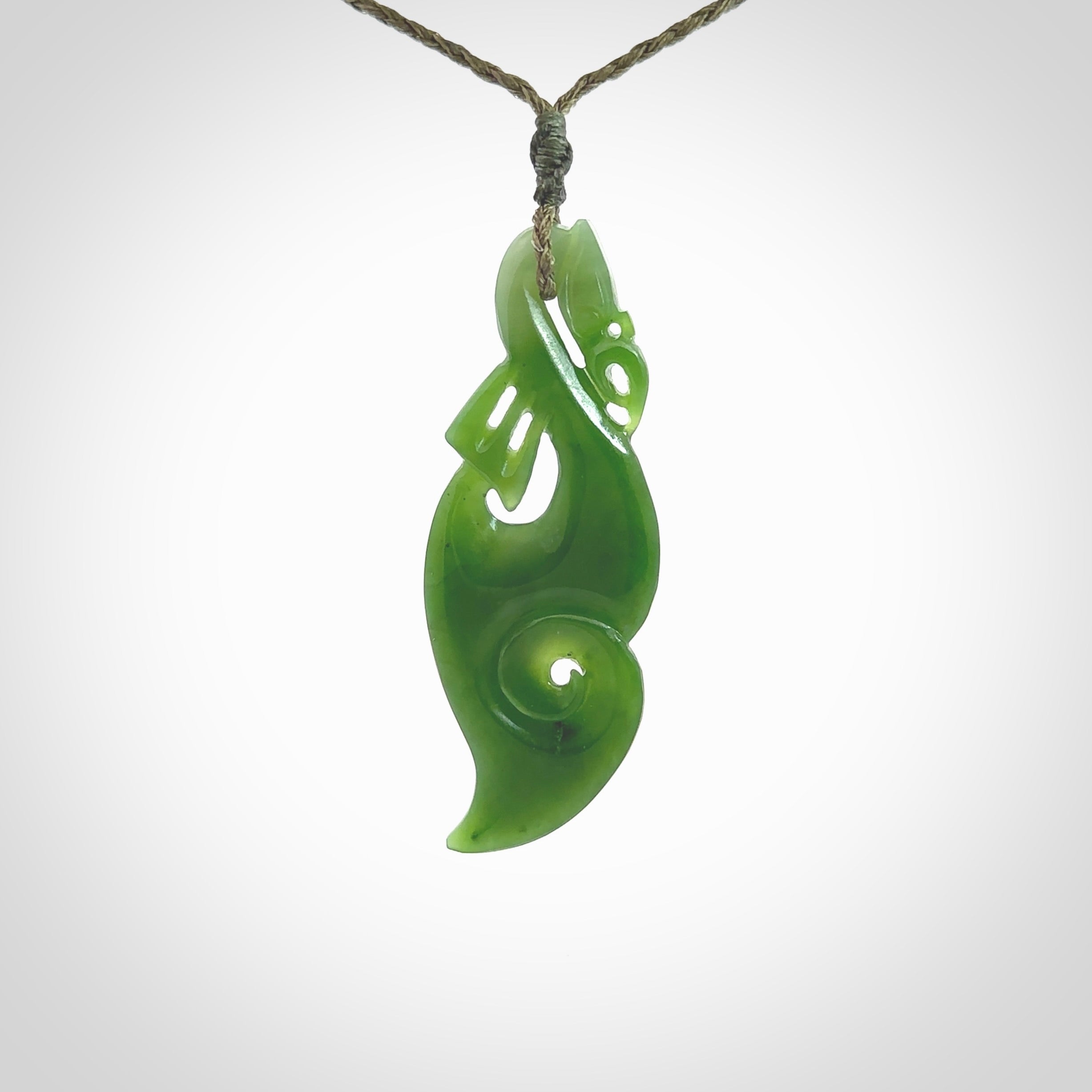 This picture shows a polished manaia pendant carved for us by Ross Crump. It is a dark matte green colour in traditional manaia design. Ross has added a koru on the bottom which elongates the carving and gives it some decorative flair. We have added a 4-Plait cord in olive green and made it length adjustable. This is a very beautiful piece of art to wear.