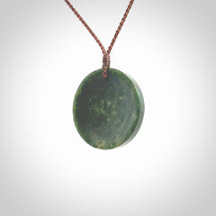 This piece is a medium sized oval round, cupped disc pendant. It was carved for us by Ric Moor from a lovely dark and varied green piece of New Zealand jade. It is suspended on a brown coloured braided cord that is length adjustable.