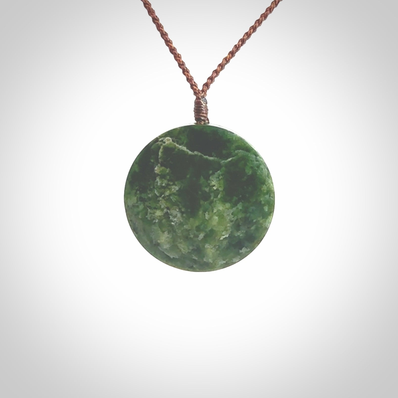 This piece is a medium sized oval round, cupped disc pendant. It was carved for us by Ric Moor from a lovely dark and varied green piece of New Zealand jade. It is suspended on a brown coloured braided cord that is length adjustable.