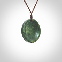 This piece is a medium sized oval round, cupped disc pendant. It was carved for us by Ric Moor from a lovely dark and varied green piece of New Zealand jade. It is suspended on a brown coloured braided cord that is length adjustable.