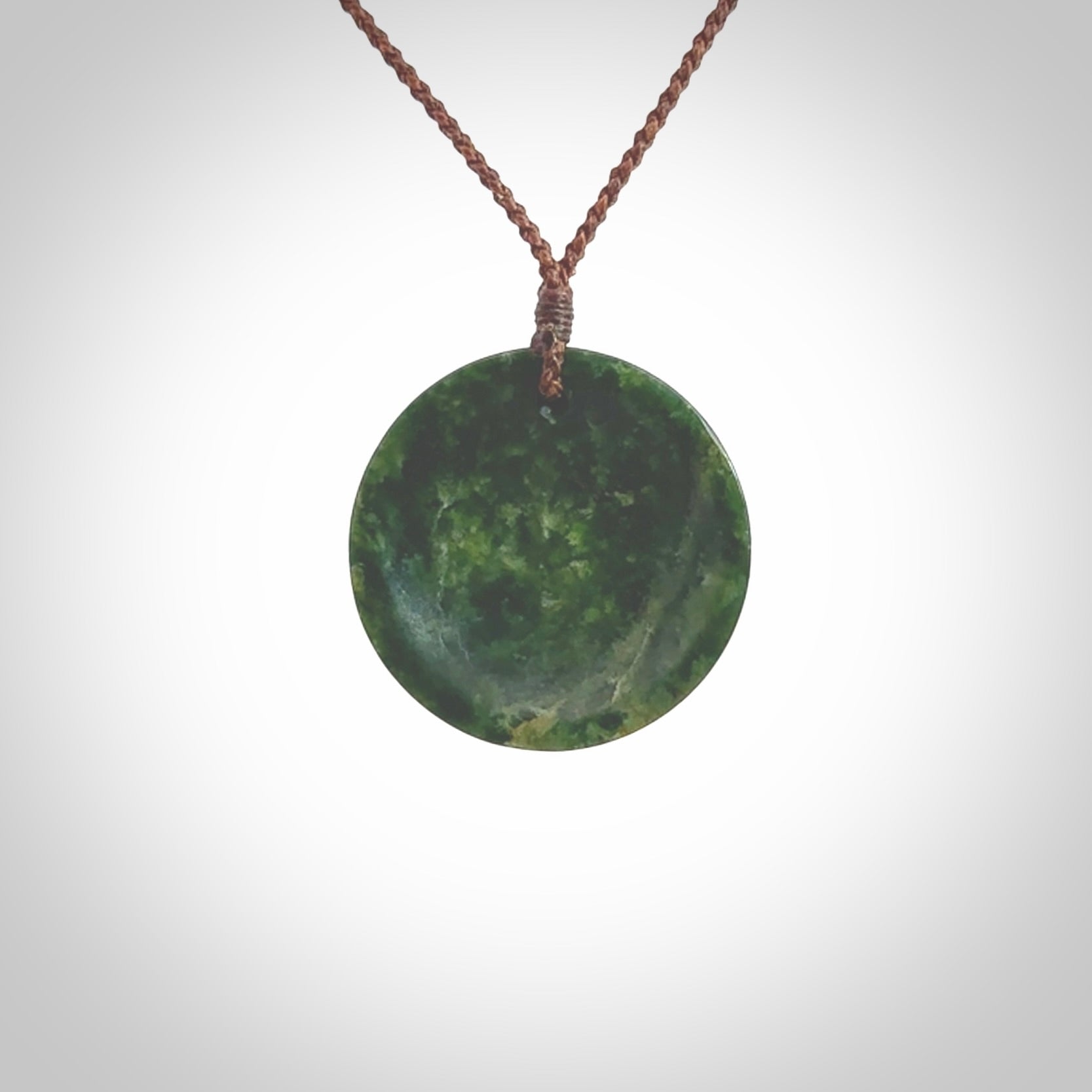 This piece is a medium sized oval round, cupped disc pendant. It was carved for us by Ric Moor from a lovely dark and varied green piece of New Zealand jade. It is suspended on a brown coloured braided cord that is length adjustable.