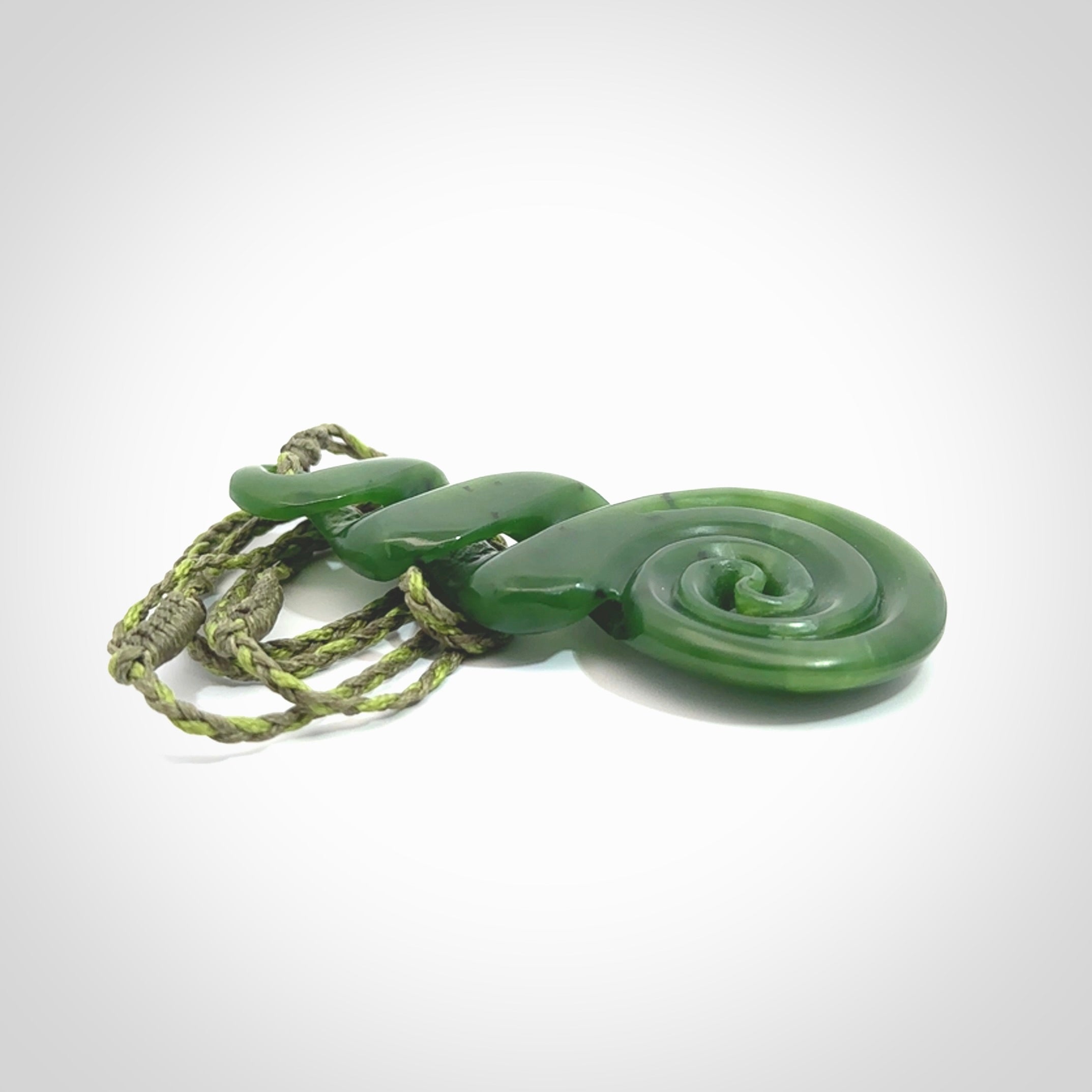 Ross Crump triple twist with koru pendant. Hand carved from rare New Zealand jade this is a beautiful pounamu pendant. The cord is 4-plait in a grass green and khaki colour and is length adjustable. It has a floret in khaki green. It is a delicate and very beautiful greenstone pendant. For sale online by NZ Pacific.