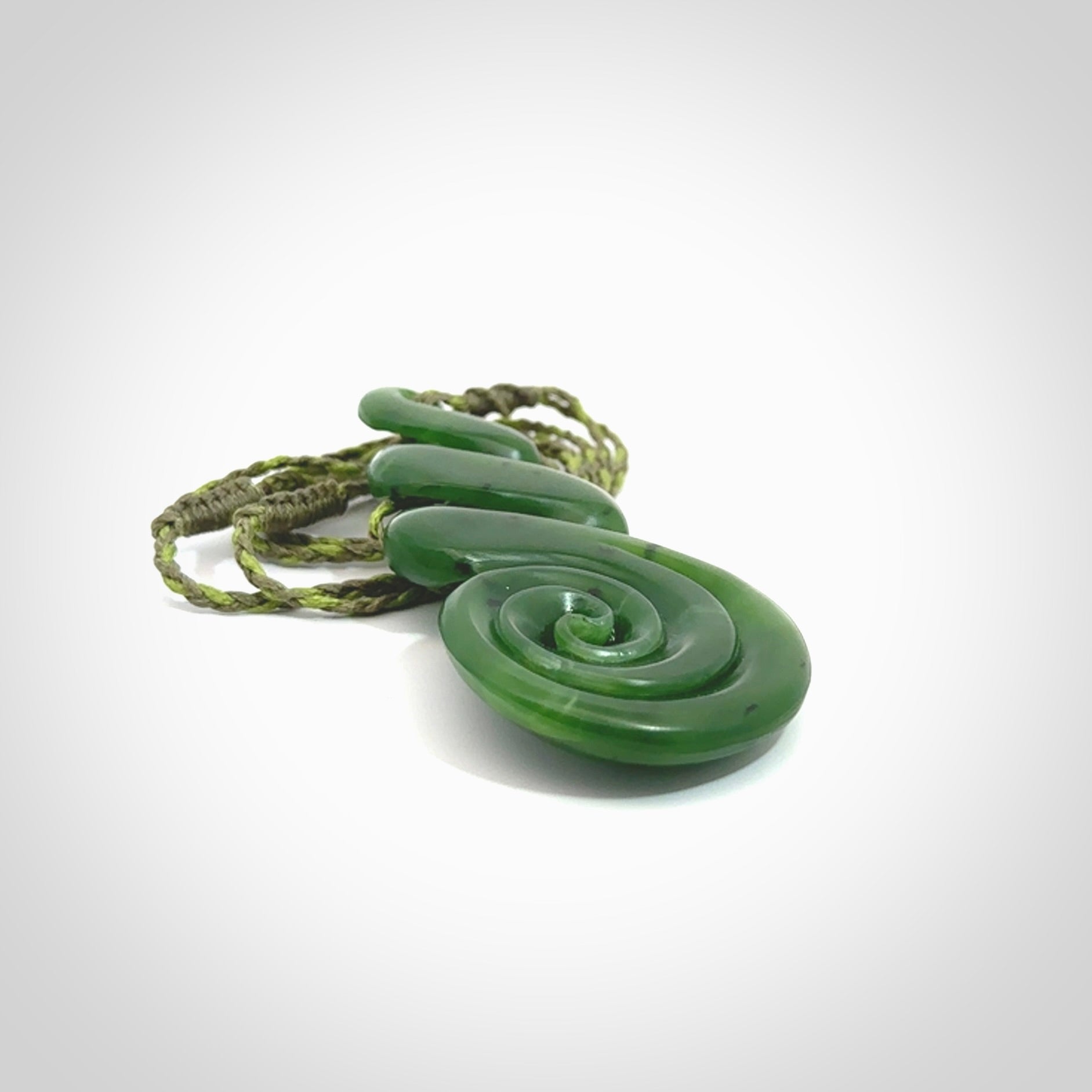 Ross Crump triple twist with koru pendant. Hand carved from rare New Zealand jade this is a beautiful pounamu pendant. The cord is 4-plait in a grass green and khaki colour and is length adjustable. It has a floret in khaki green. It is a delicate and very beautiful greenstone pendant. For sale online by NZ Pacific.