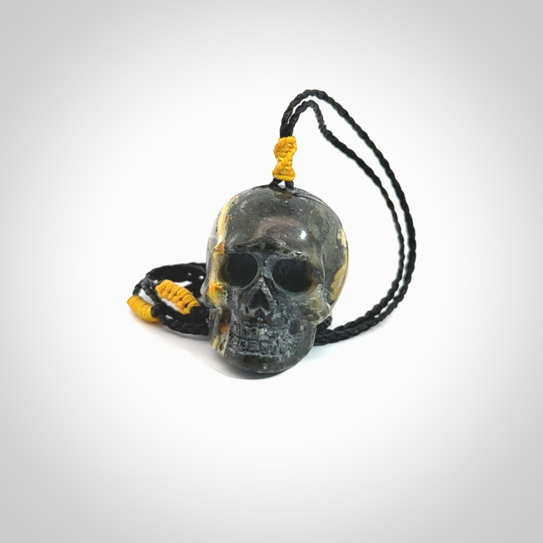 This picture shows a large sized Bumblebee Jasper stone skull pendant that we have hand carved. It is polished to a soft shine and is a very striking piece. The cord is a black colour and has a small yellow floret binding just above the pendant.