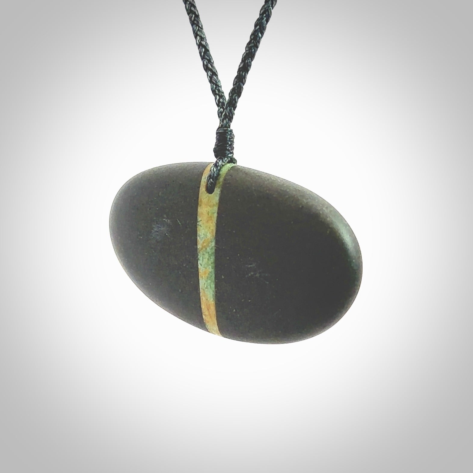 A drop pendant made from Onewa Stone - New Zealand Greywacke - with a fine Pounamu insert spliced into the middle. Hand carved stone drop with New Zealand Jade centre. Provided with an adjustable charcoal grey cord and packaged in a woven kete pouch.