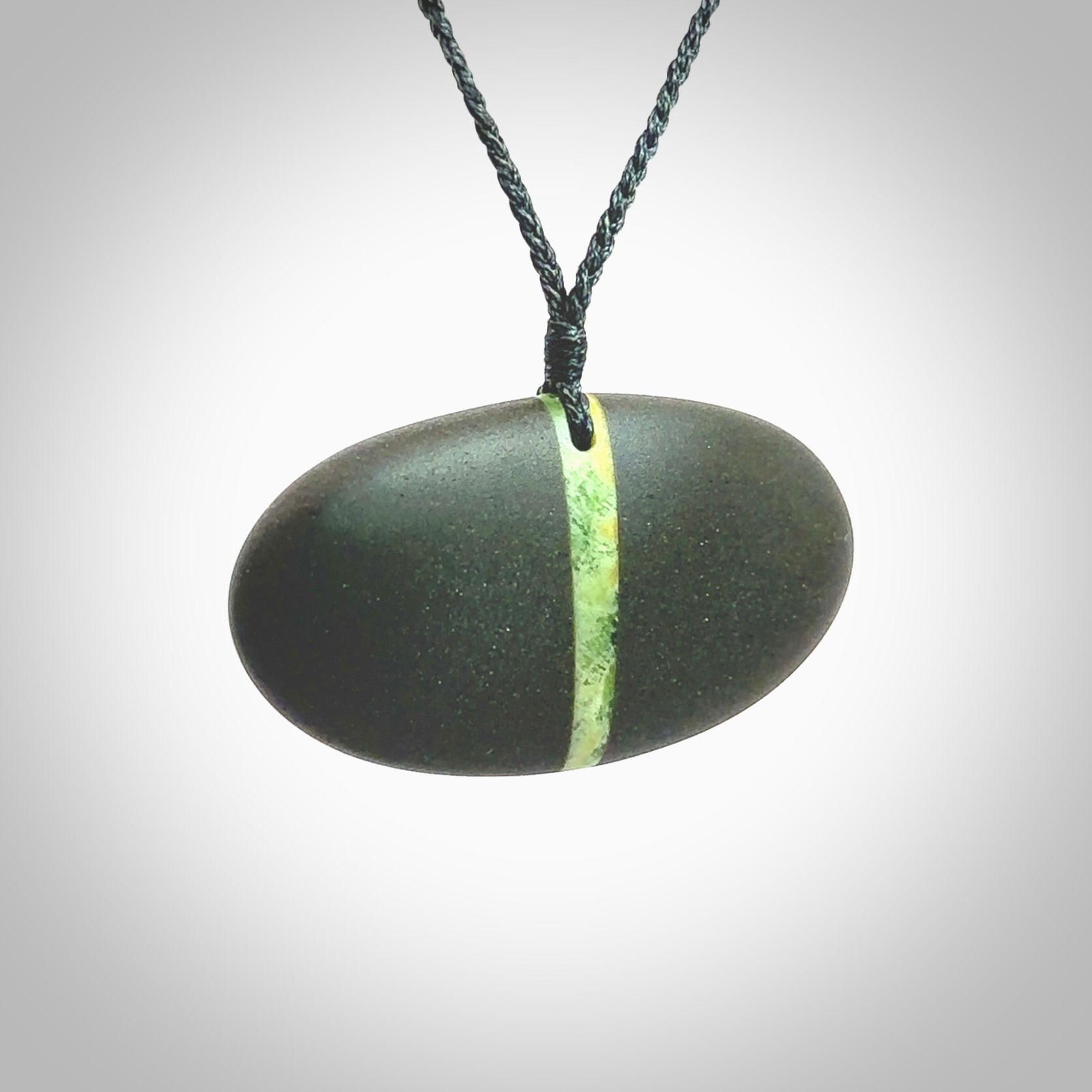 A drop pendant made from Onewa Stone - New Zealand Greywacke - with a fine Pounamu insert spliced into the middle. Hand carved stone drop with New Zealand Jade centre. Provided with an adjustable charcoal grey cord and packaged in a woven kete pouch.