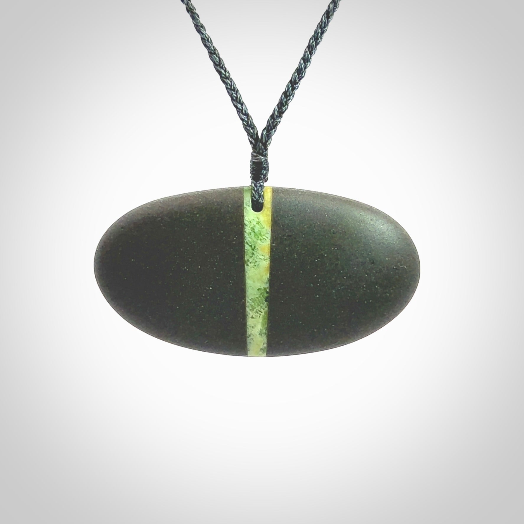 A drop pendant made from Onewa Stone - New Zealand Greywacke - with a fine Pounamu insert spliced into the middle. Hand carved stone drop with New Zealand Jade centre. Provided with an adjustable charcoal grey cord and packaged in a woven kete pouch.