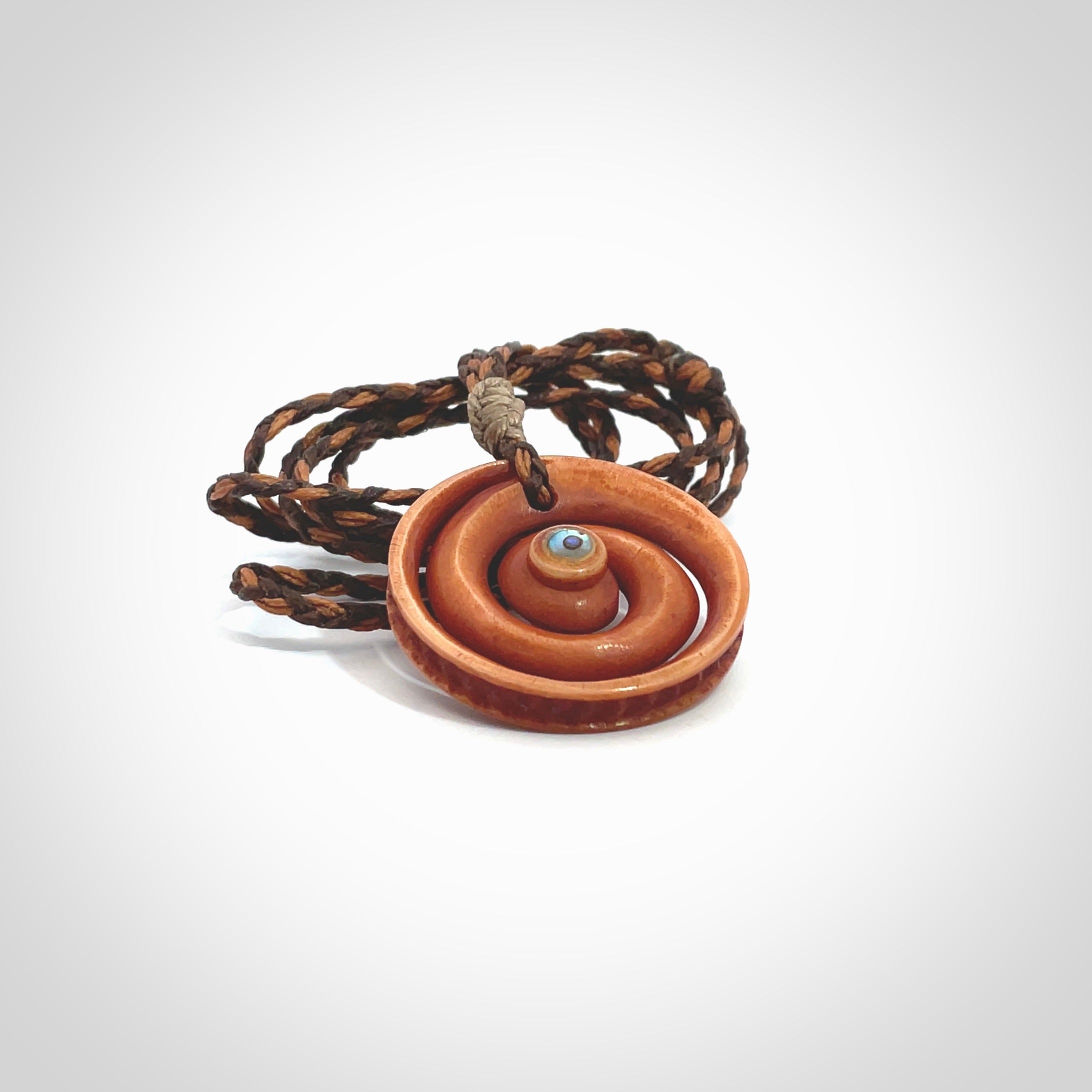 A hand carved and intricate koru pendant made for us by Yuri Terenyi. This is a beautiful little piece and is emblematic of the well known and loved Koru design. It is carved from stained bone in a hollowed, koru spiral shape with paua shell inlay in the middle of the koru. It is suspended from a gingernut brown/wapiti brown cord with a stardust floret and the necklace is adjustable.