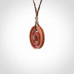 A hand carved and intricate koru pendant made for us by Yuri Terenyi. This is a beautiful little piece and is emblematic of the well known and loved Koru design. It is carved from stained bone in a hollowed, koru spiral shape with paua shell inlay in the middle of the koru. It is suspended from a gingernut brown/wapiti brown cord with a stardust floret and the necklace is adjustable.