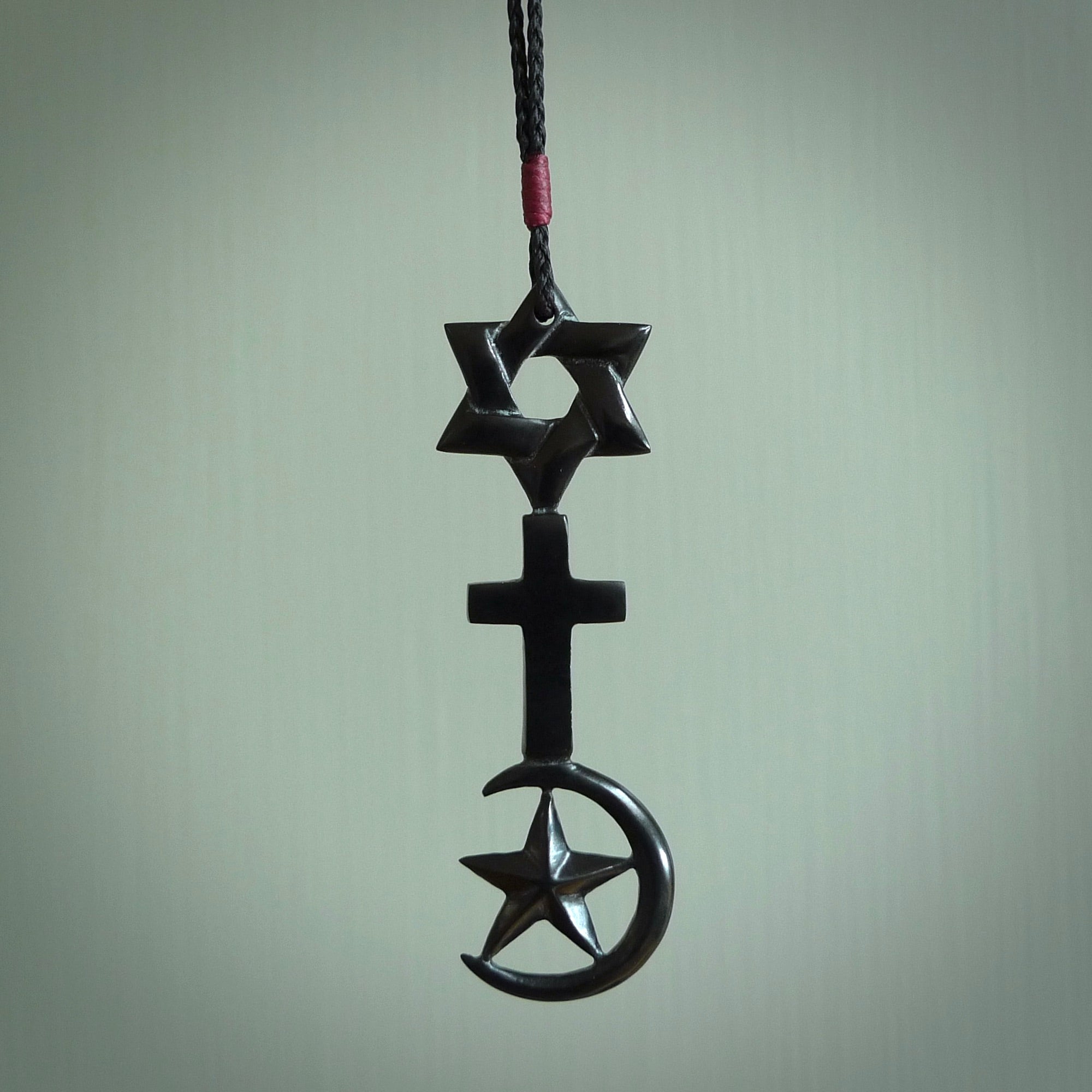 This pendant is a series of religious symbols carved in one piece of black jade. The top is a star of david, next is a christian cross and at the bottom is the crescent of islam. It is suspended on a fine, plaited black cord which is length adjustable.